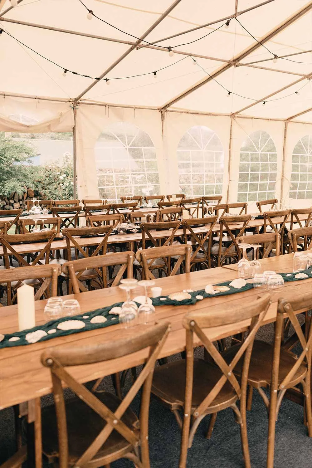 A large outdoor tented dining area with long wooden tables and cross-back chairs, decorated with seashells, candles, and glassware for an event.