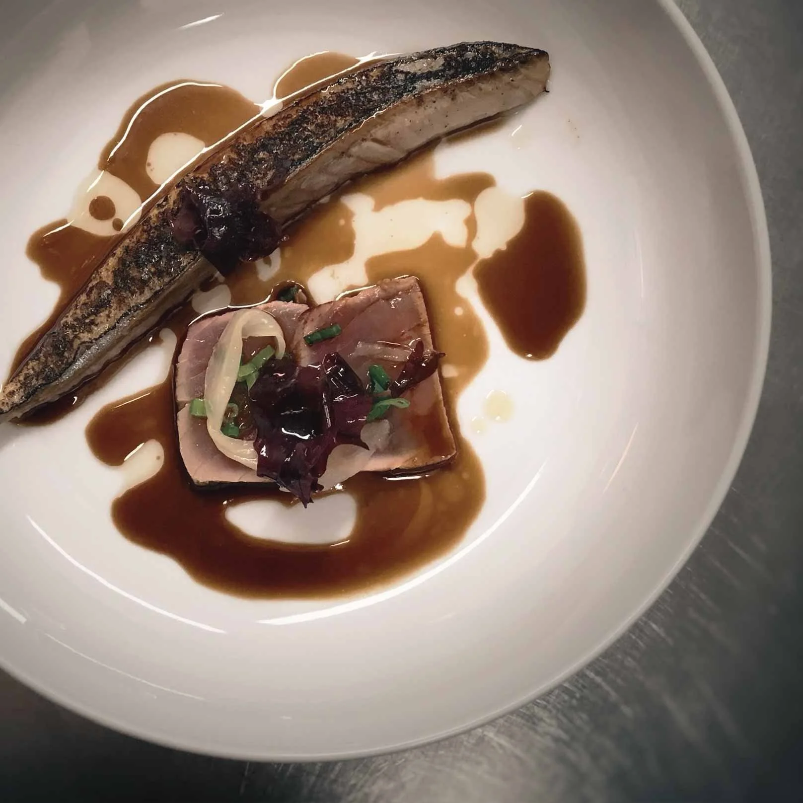 A plated dish featuring a fillet of grilled fish with dark crispy skin, served with a sauce drizzled over it and a piece of rolled meat topped with a dark sauce, garnished with chopped green onions.