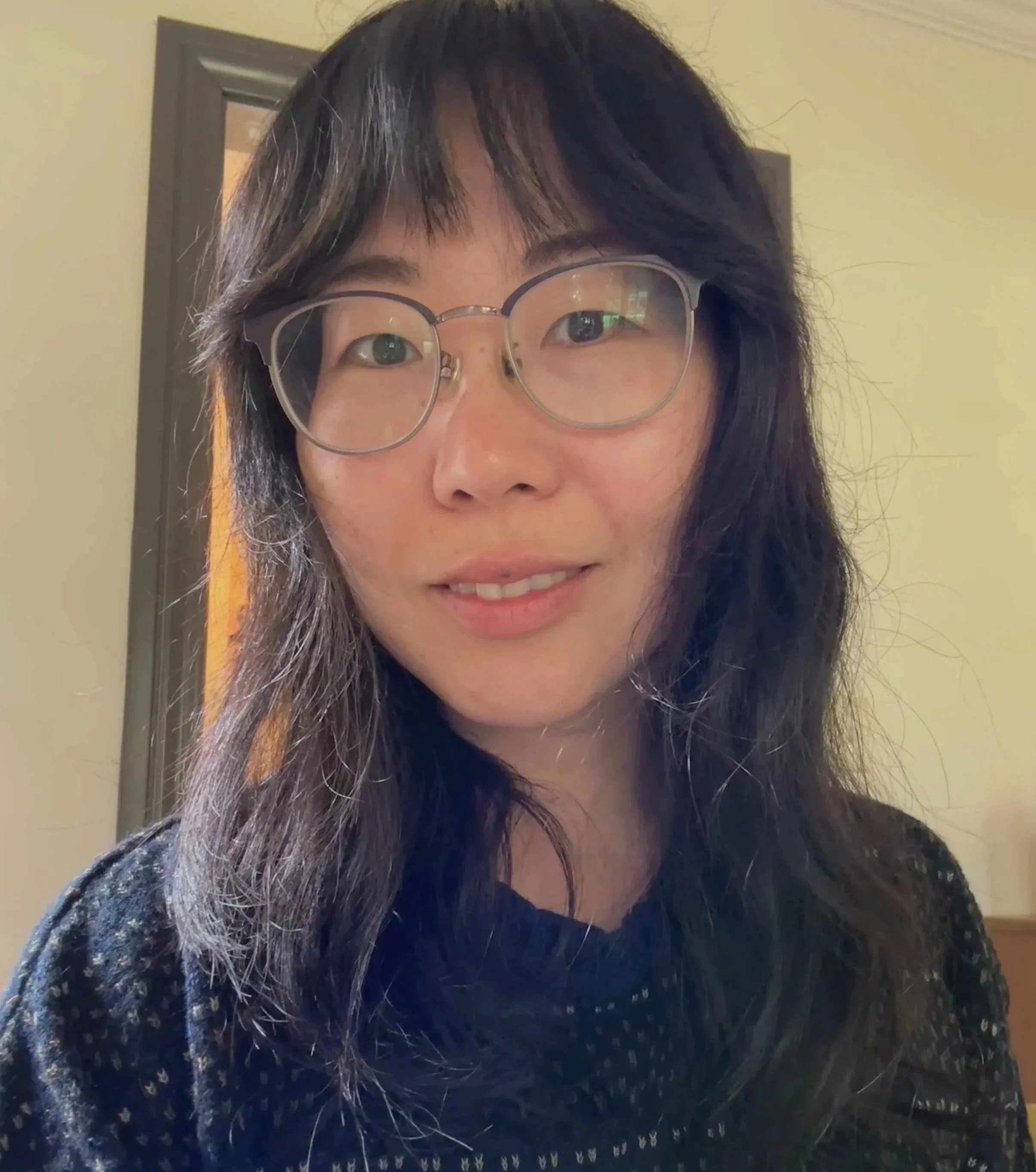 Close-up of a woman's face showing her eyes, glasses, and hair, with a light-colored wall and a door in the background.