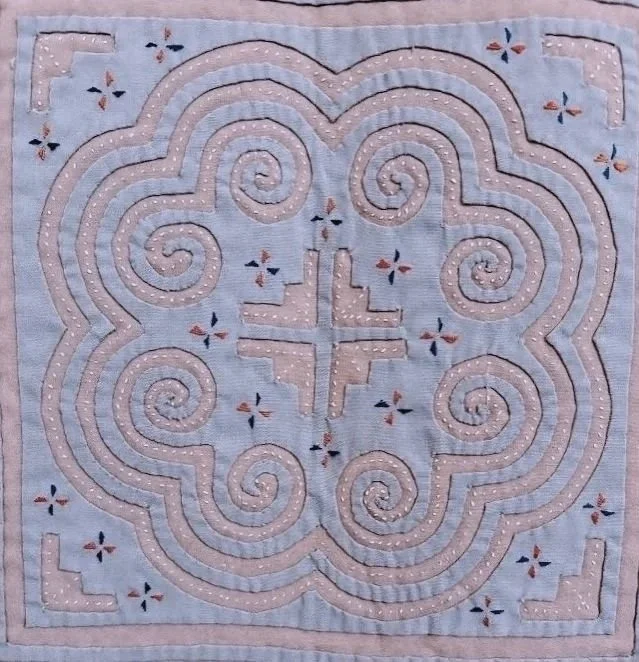 A fabric piece with intricate embroidery featuring swirling patterns, a central cross-like design, and small decorative motifs around the edges, all with pastel colors.