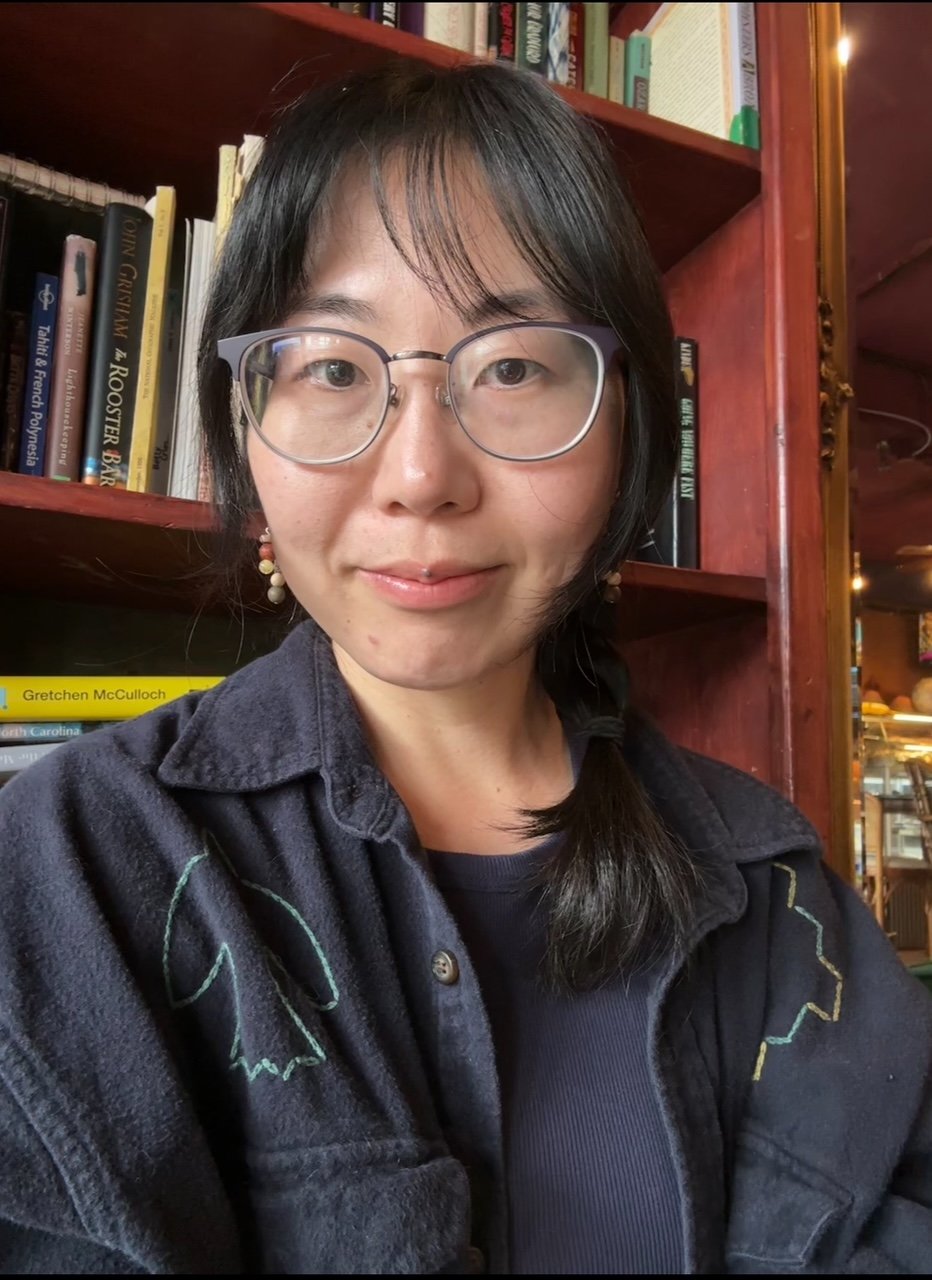A woman with short black hair, glasses, and earrings, taking a selfie in a bookstore or library.