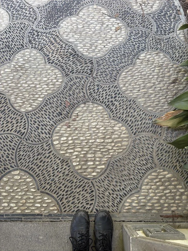 A decorative sidewalk with a pattern of grey and white stones, viewed from above, with black shoes visible at the bottom edge.