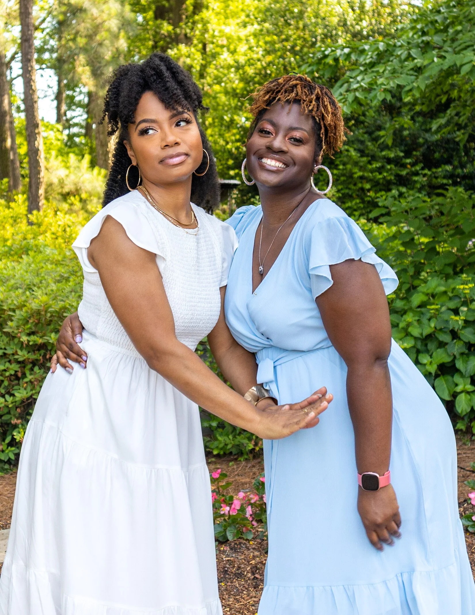 Two women outdoors, smiling and embracing, standing in front of green foliage and trees.