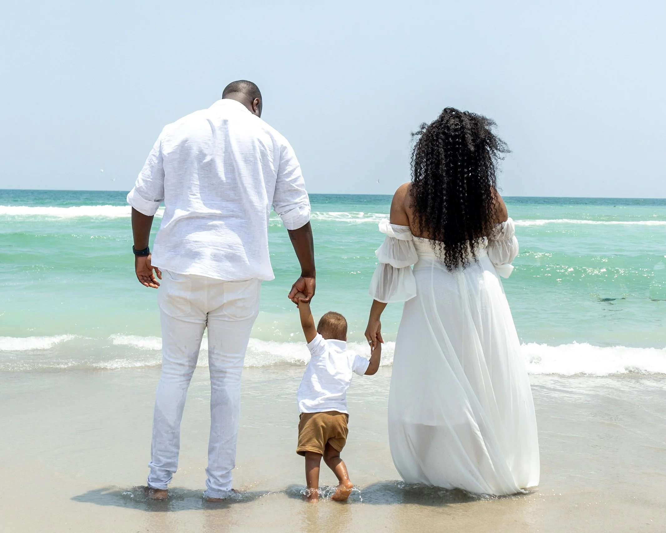 A family of four holding hands, walking into the ocean at the beach with blue water and a light sky.