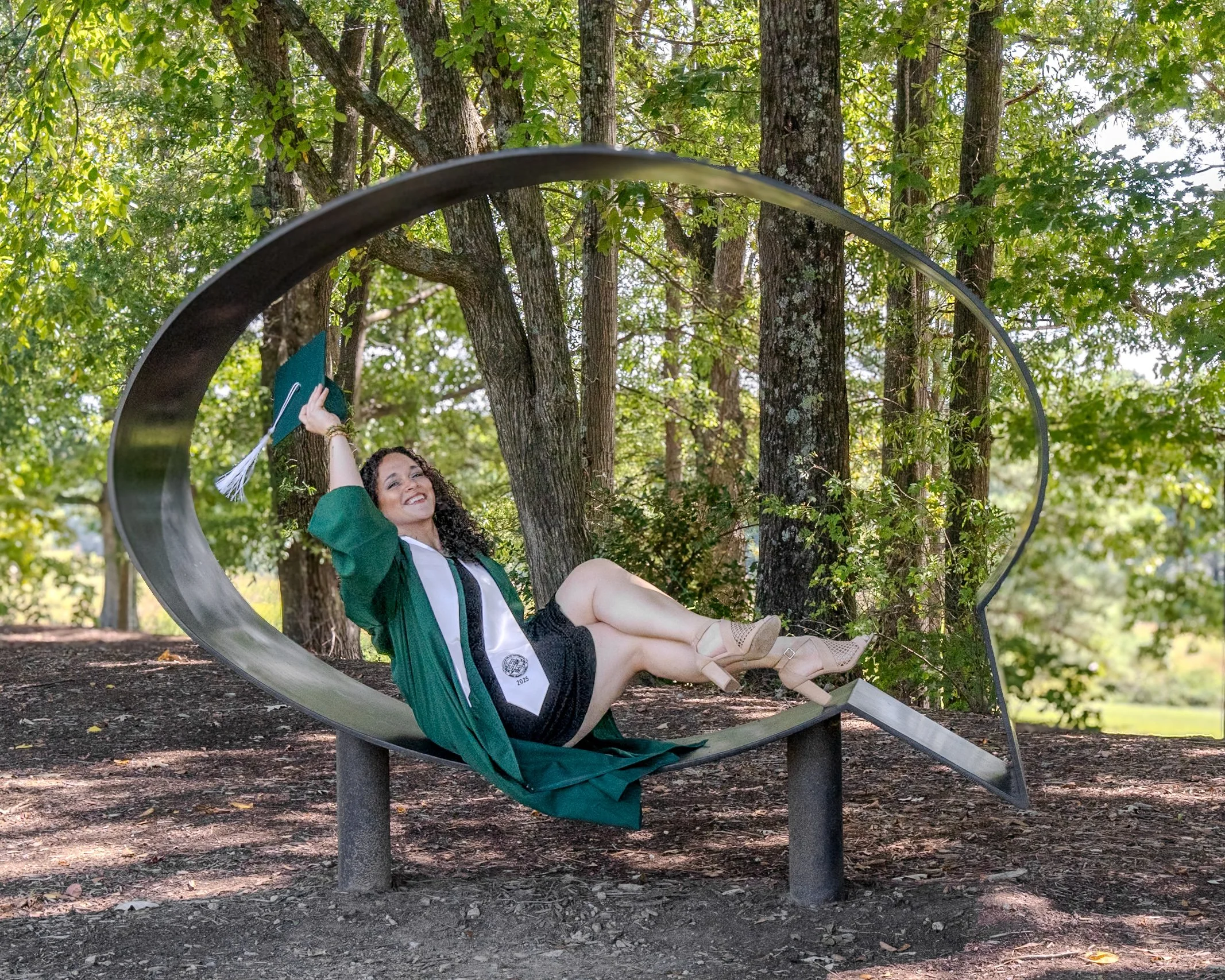 A young woman with curly hair smiling and sitting on a park bench with a metallic circular sculpture behind her. Graduation photoshoot. Art lover. Content Creation.
