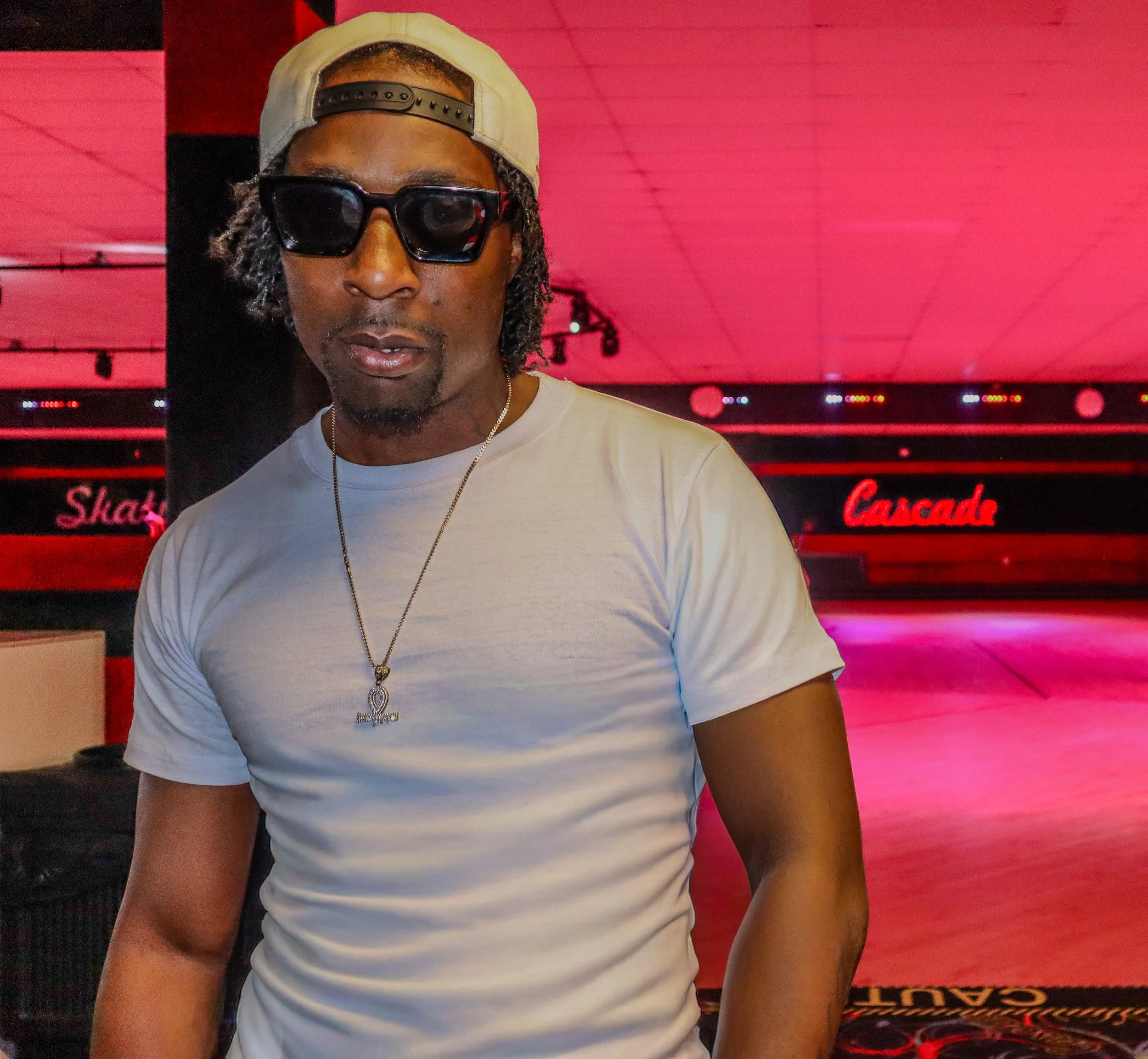 Close-up of a person wearing sunglasses, a beige cap backward, a white T-shirt, a silver chain with a pendant, Music Artist/rapper at Cascade Skating Rink. Atlanta, Georgia. 