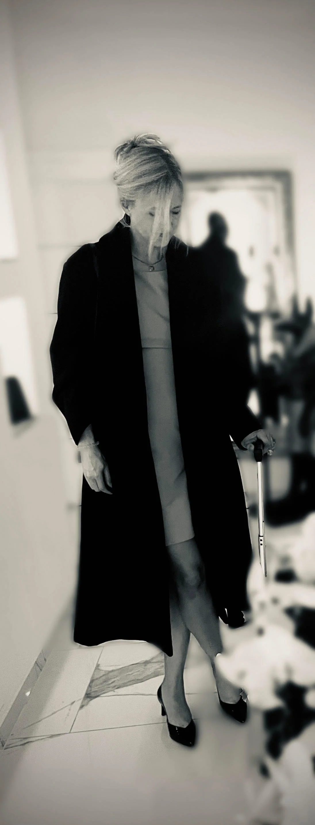 Frau mit blonden Haaren, schwarzem Mantel, grauem Kleid, schwarzen High Heels und Kabinenkoffer, im Innenraum, unscharfer Hintergrund, schwarz-weiß Foto.