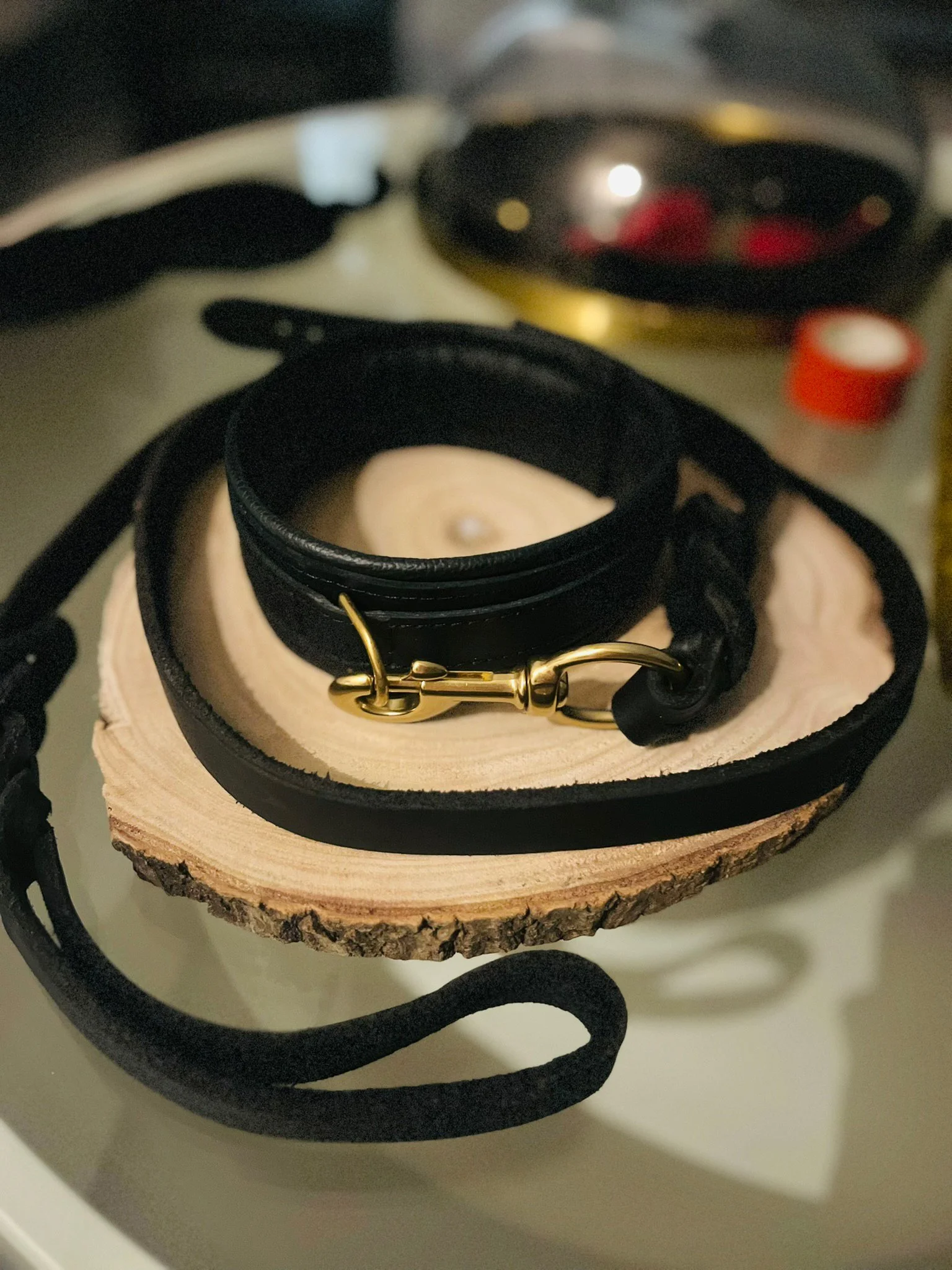 Ein Lederhalsband mit goldenem Karabiner liegt auf einem Holzstück. Im Hintergrund sind Unsicherheiten, die auf ein dunkles, vage Objekt hinweisen.