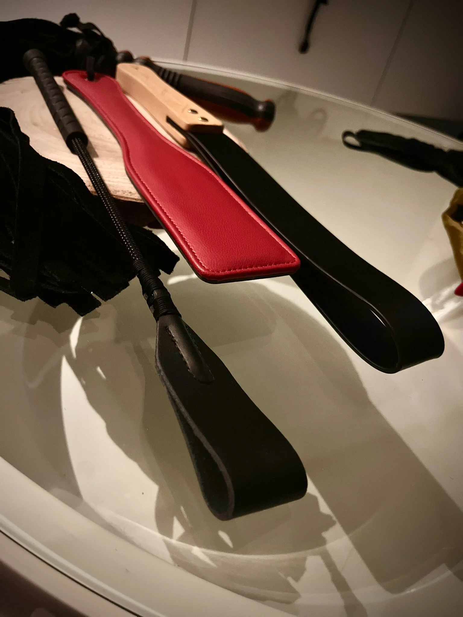 Eine gläserne Tischplatte, darauf befinden sich mehrere Gegenstände, darunter ein schwarz-rotes Lederpaddel, eine schwarze Gerte mit Klatscher, ein Lederriemenpaddel mit hellem Holzgriff und andere Schlagwerkzeuge