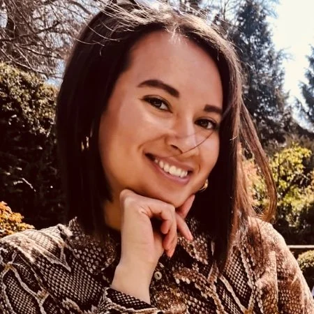 A woman smiling outdoors with trees in the background, wearing a patterned shirt.