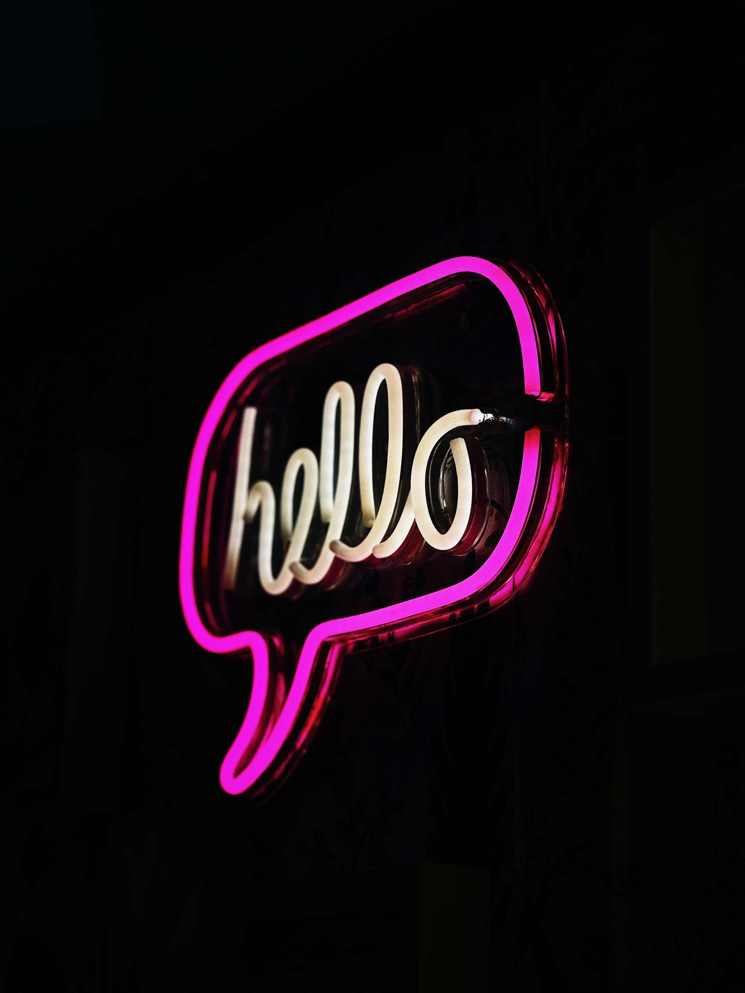 Pink and white neon sign in the shape of a speech bubble with the word 'hello' inside, against a black background.