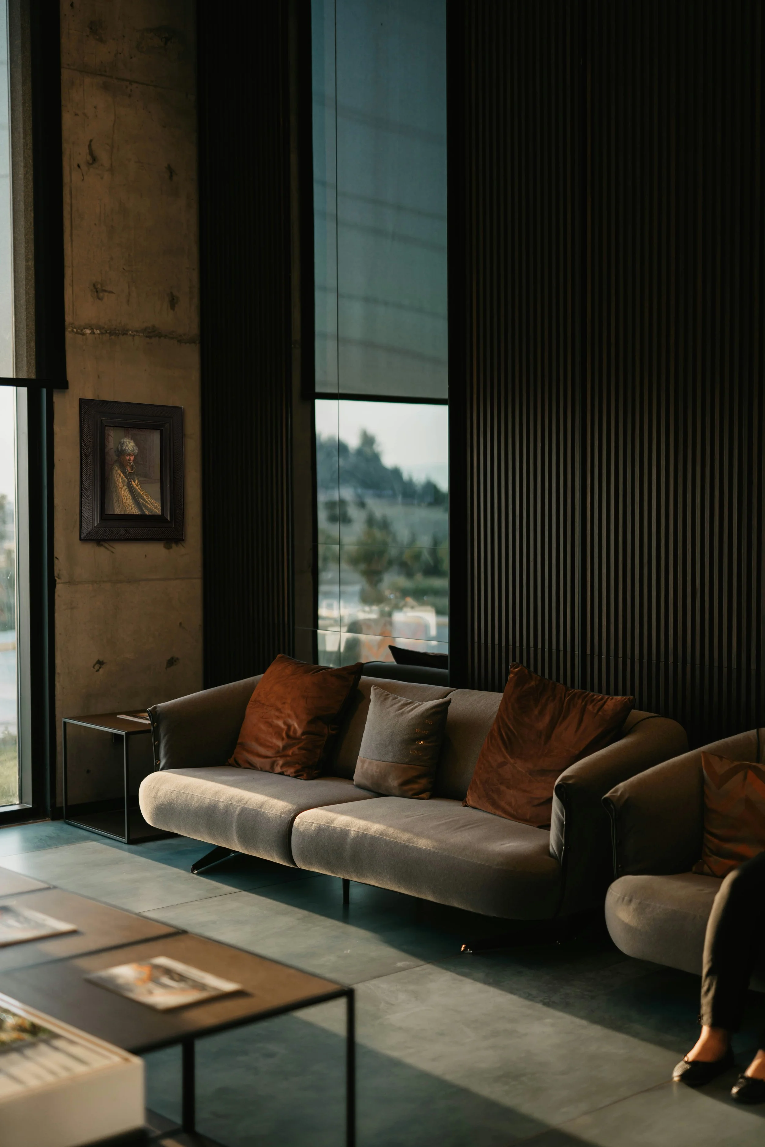 Modern dark toned living room with a framed Goldie painting on the wall next to a large window.
