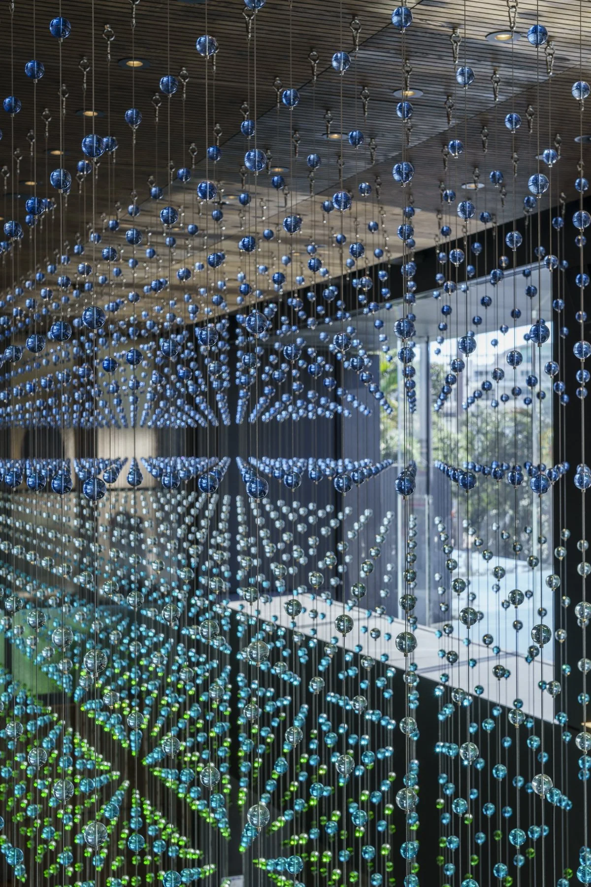 A large artistic installation by Nike Savvas made of hanging blue, green, and clear glass balls with wires, creating a cascading, wave-like pattern inside the InterContinental Hotel in Auckland