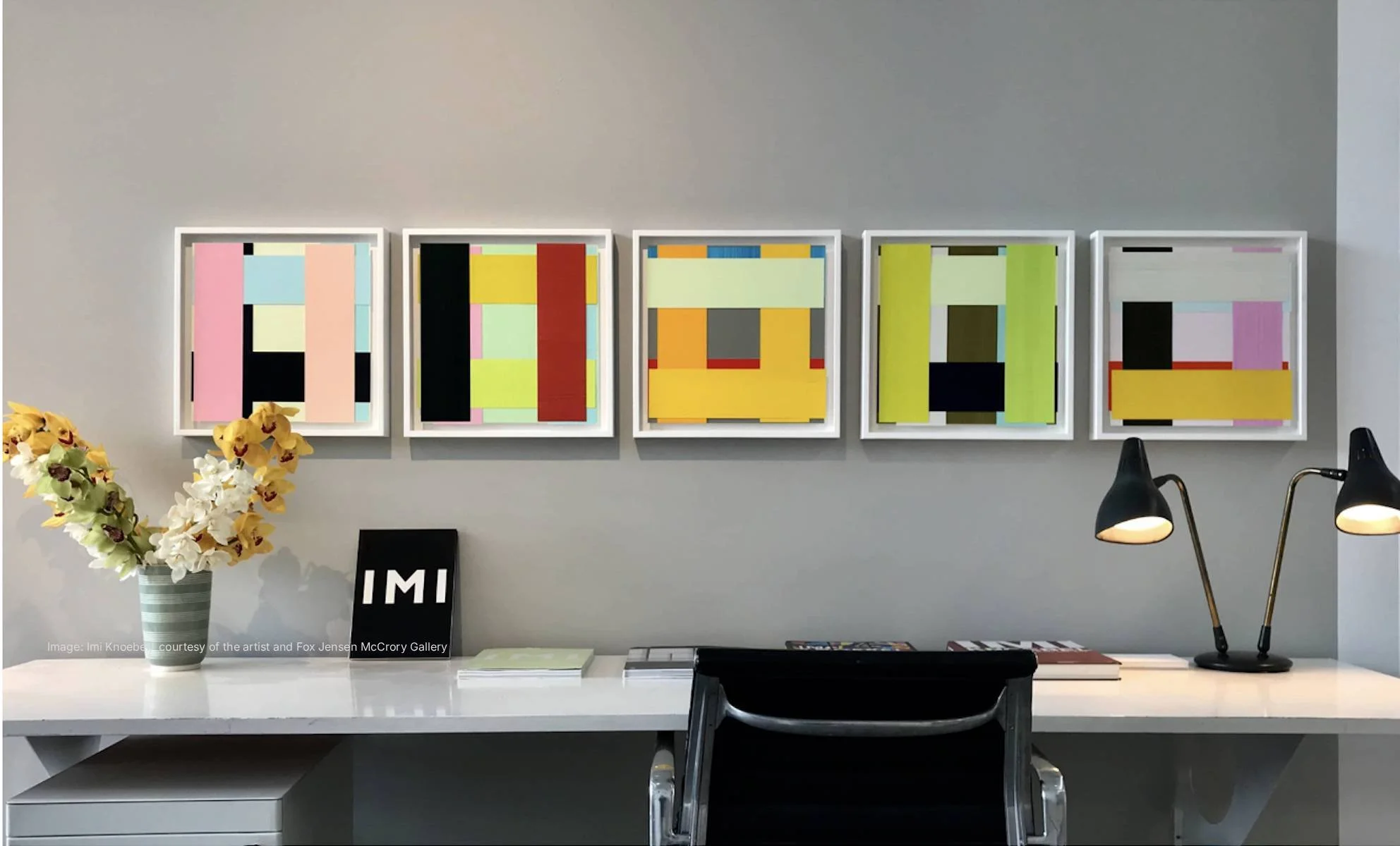 A modern art gallery display with five abstract, colorful framed artworks on a light gray wall by Imi Knoebel, a white table in front with a vase of white and yellow orchids, black letter art reading 'IMI,' and two black desk lamps.