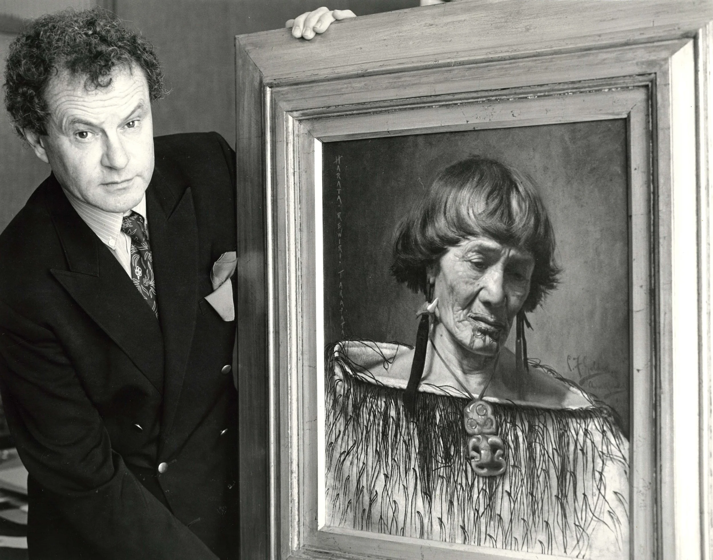 Black and white photograph of Peter Webb holding up a framed Goldie painting. 