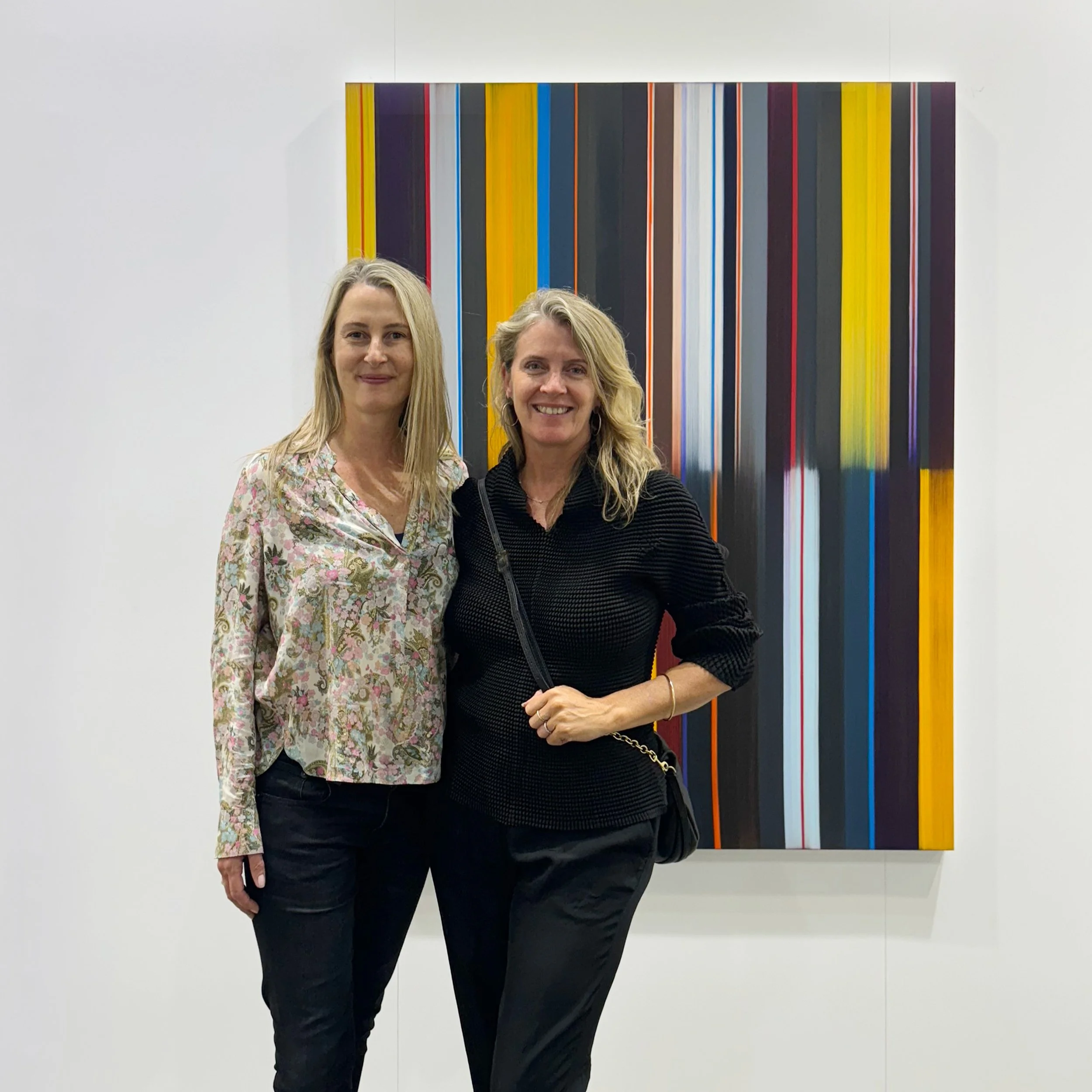 Sophie Coupland and Dr. Emily Cormack standing in front of a modern colourful striped art piece on a white wall.