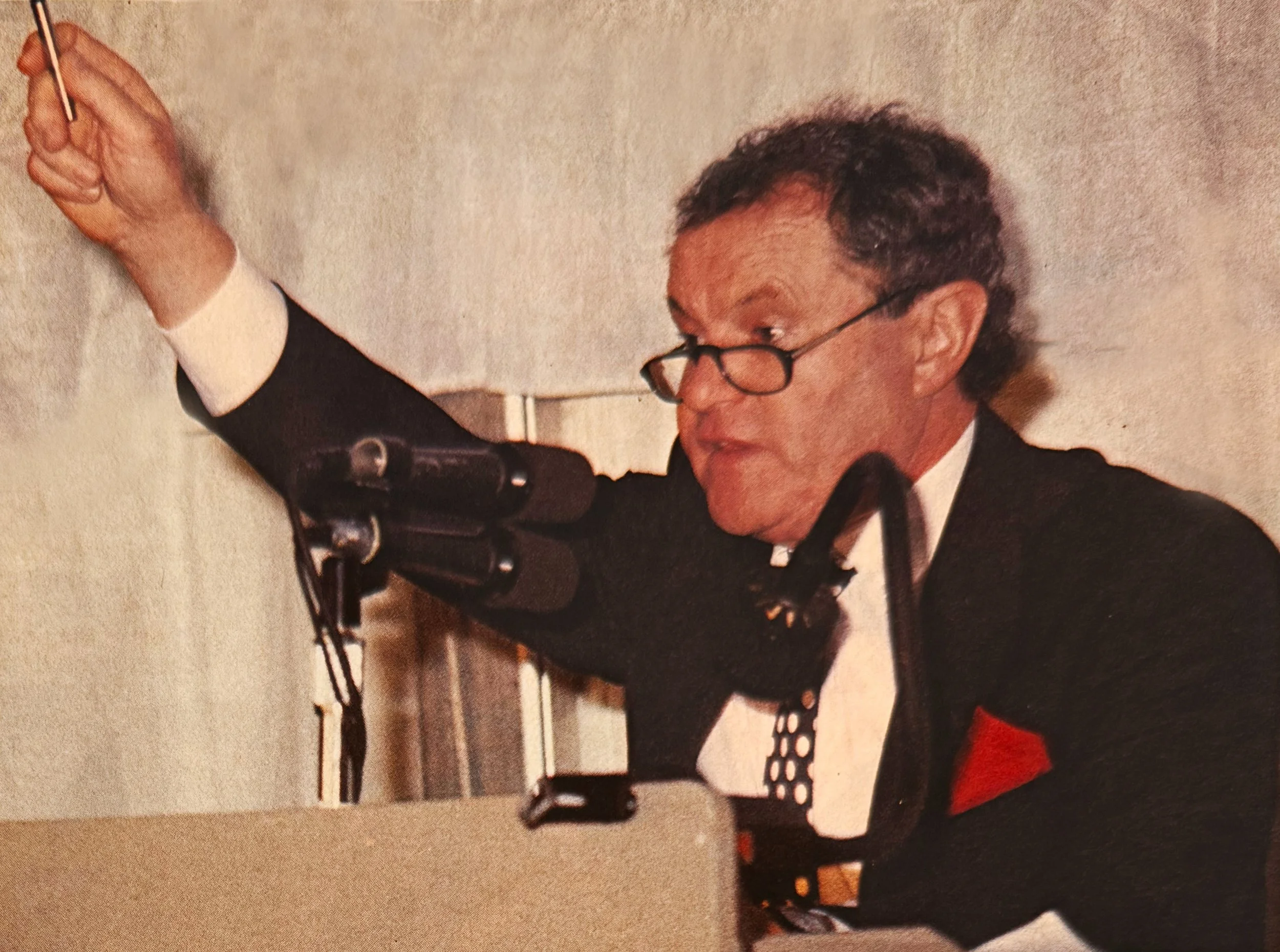 Peter Webb in a black tuxedo with a red pocket square is speaking passionately at a auctioneers podium with a microphone, raising his right hand with a pointed finger gesture, against a plain background.