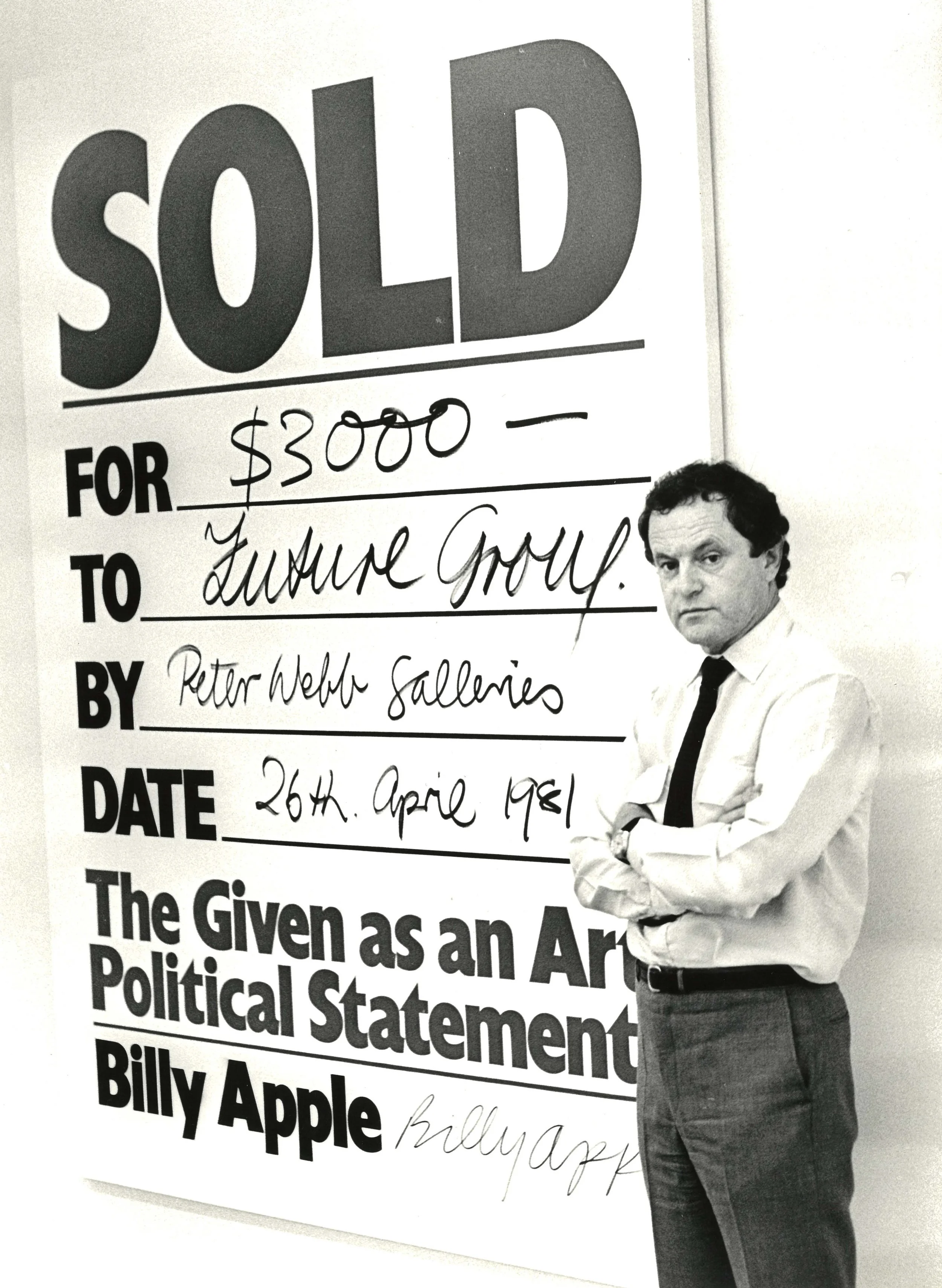 Black and white photo of Peter Webb in a white shirt and dark tie standing with arms crossed next to a large poster with the word 'SOLD' at the top and handwritten details about a sale.