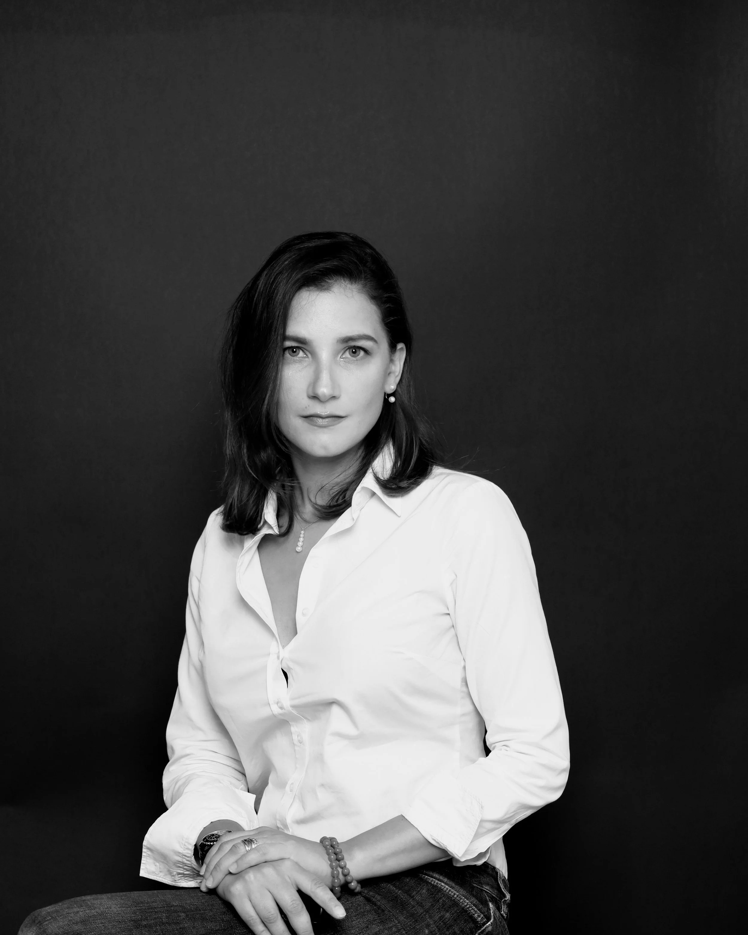 Black and white portrait of a woman with dark hair, wearing a white button-up shirt and jewelry, sitting with her hands crossed on her lap against a dark background.
