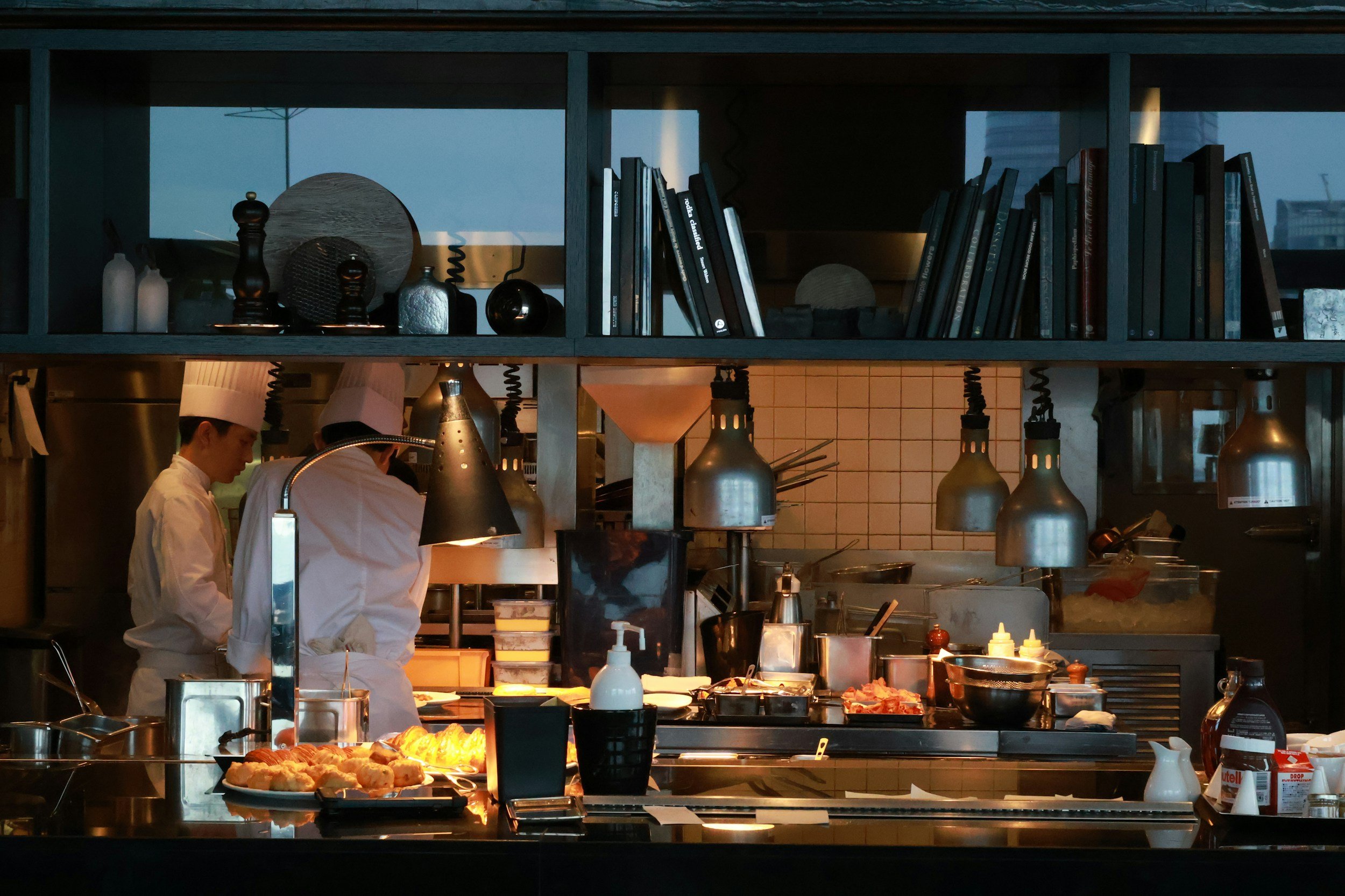 A busy kitchen scene with two chefs preparing food, featuring shelves with books and decorative items, hanging lamps, and a counter with various dishes and condiments.