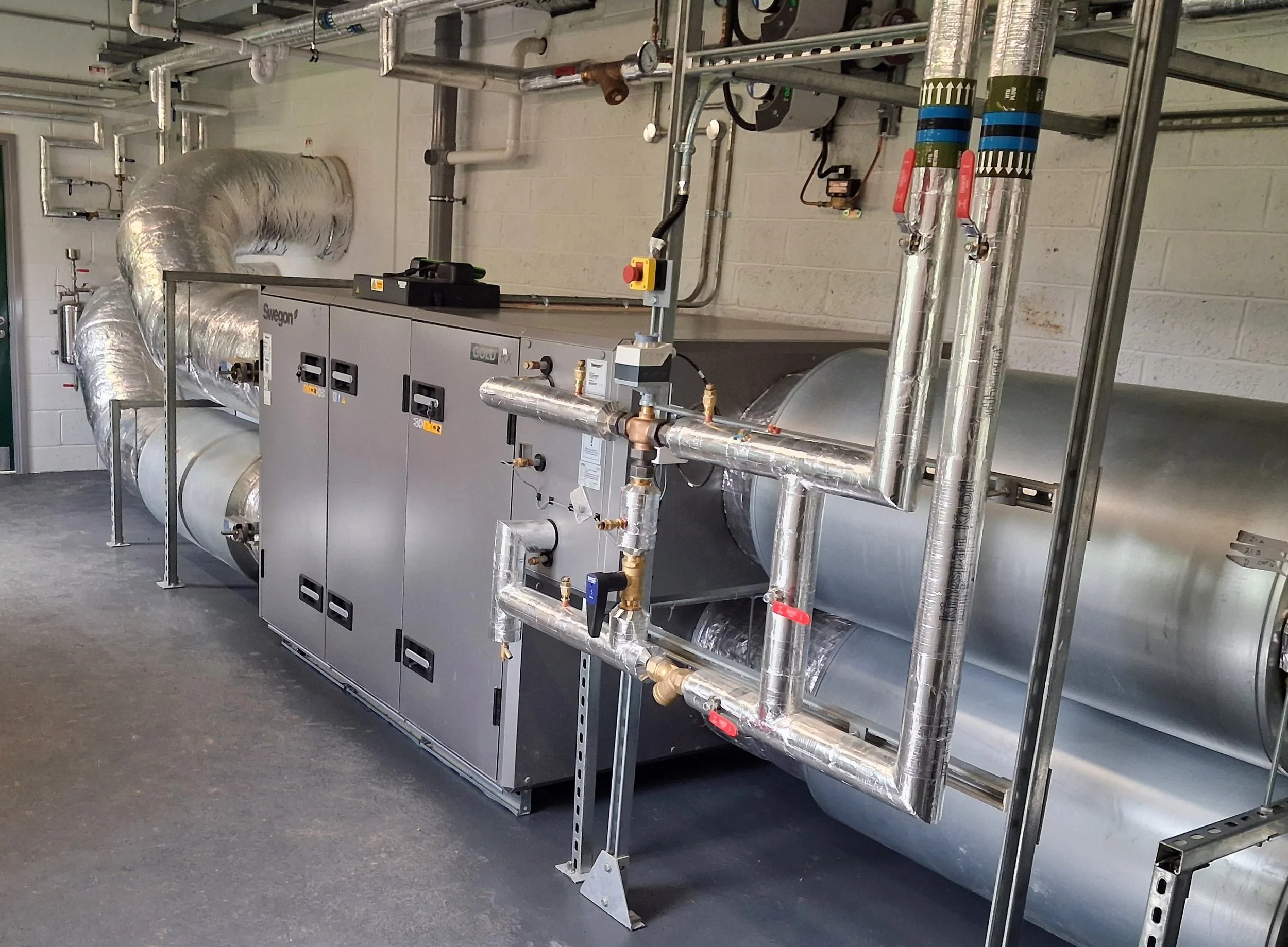 Industrial HVAC system with various metal and insulated pipes, gauges, valves, and control panels mounted on a metal frame inside a utility room.