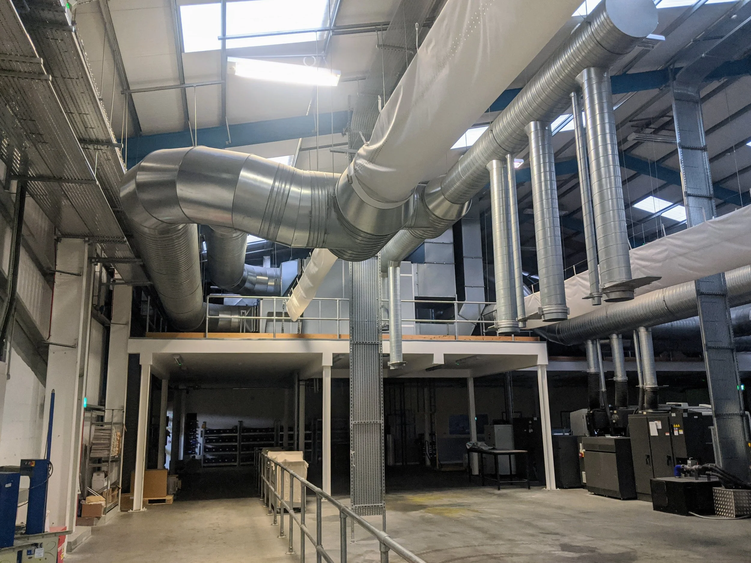 Interior view of an industrial facility with large silver duct pipes running across the ceiling, metal scaffolding, and machinery along the walls.