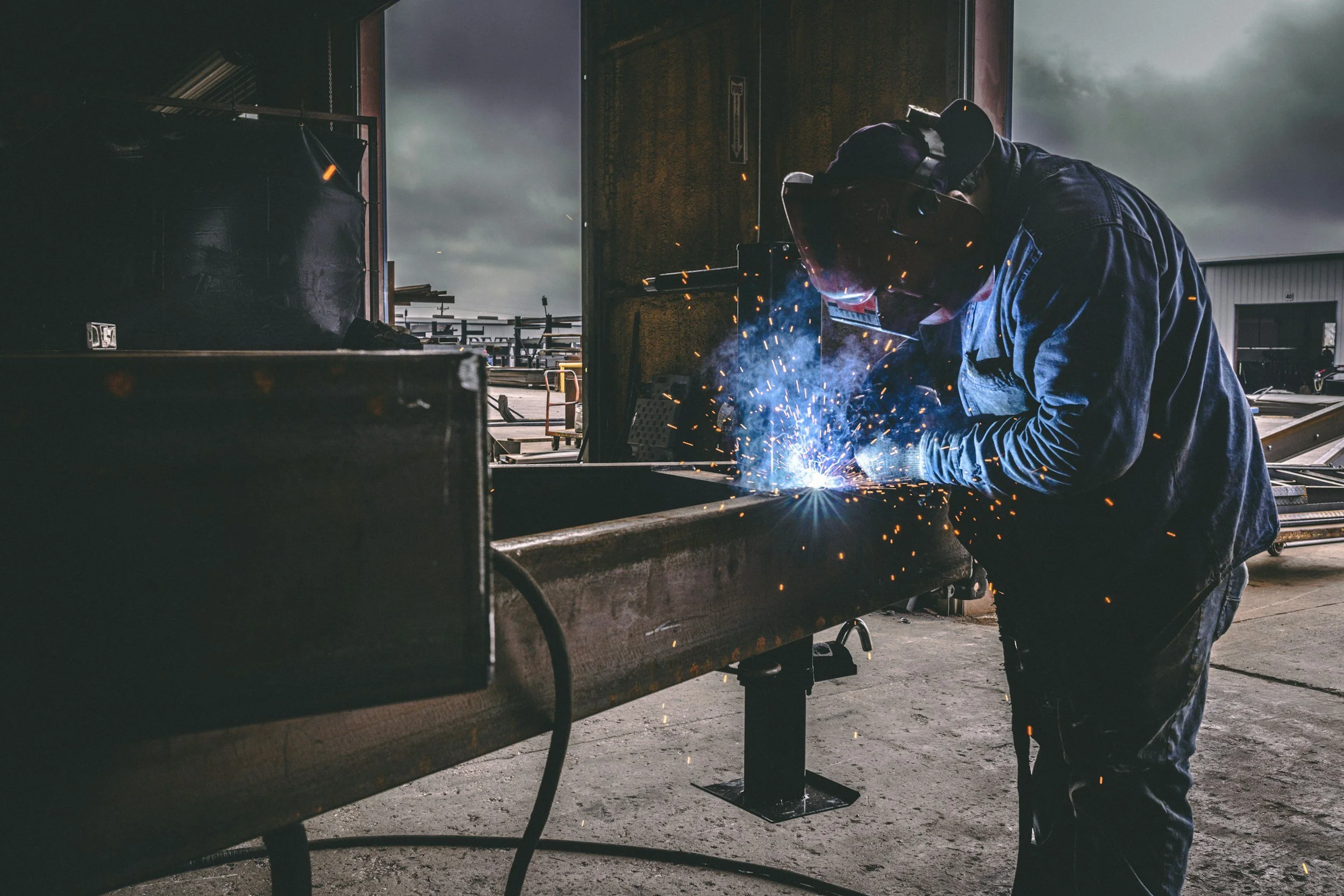 A worker welding metal on a workbench inside a workshop, with sparks flying from the welding process.