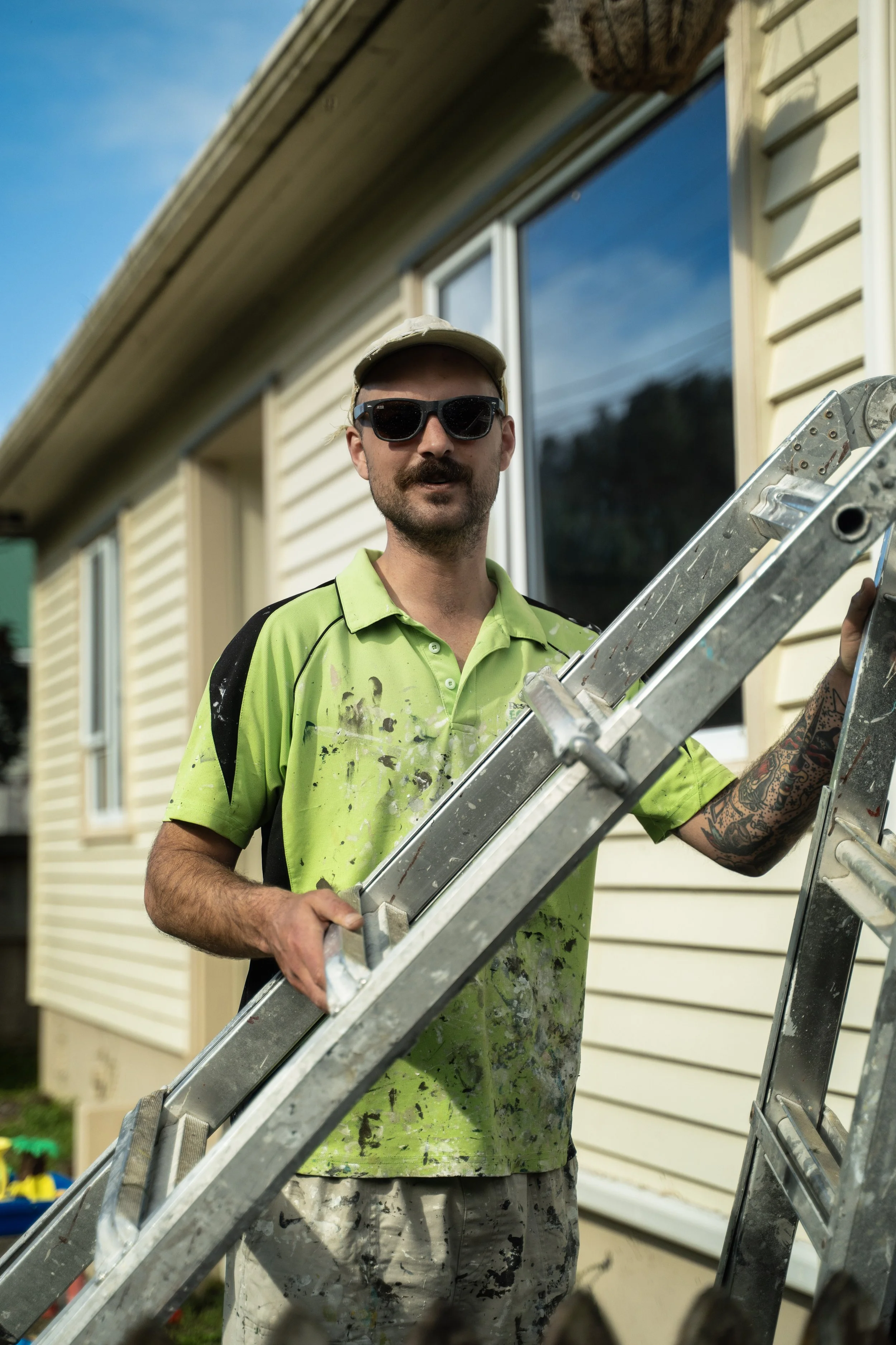 A man with a mustache and tattoos wearing safety sunglasses, a beige cap, a bright green paint-splattered shirt, and gray camouflage pants holding a ladder outside a house with light yellow siding and a large window, during daytime.