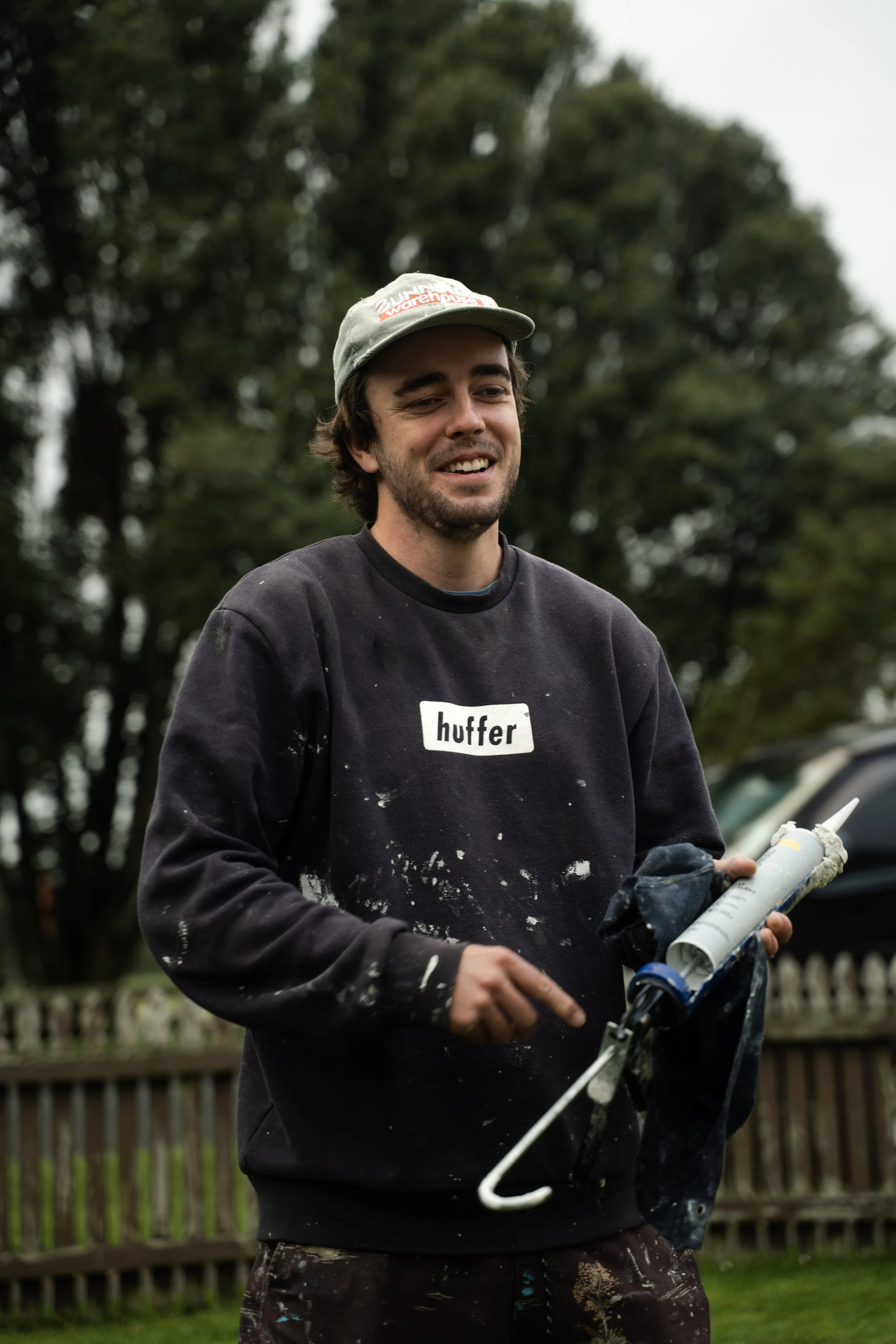A man wearing a gray cap and a black sweatshirt with the word 'huffer' on it, holding a paint roller and a paint bucket outdoors.