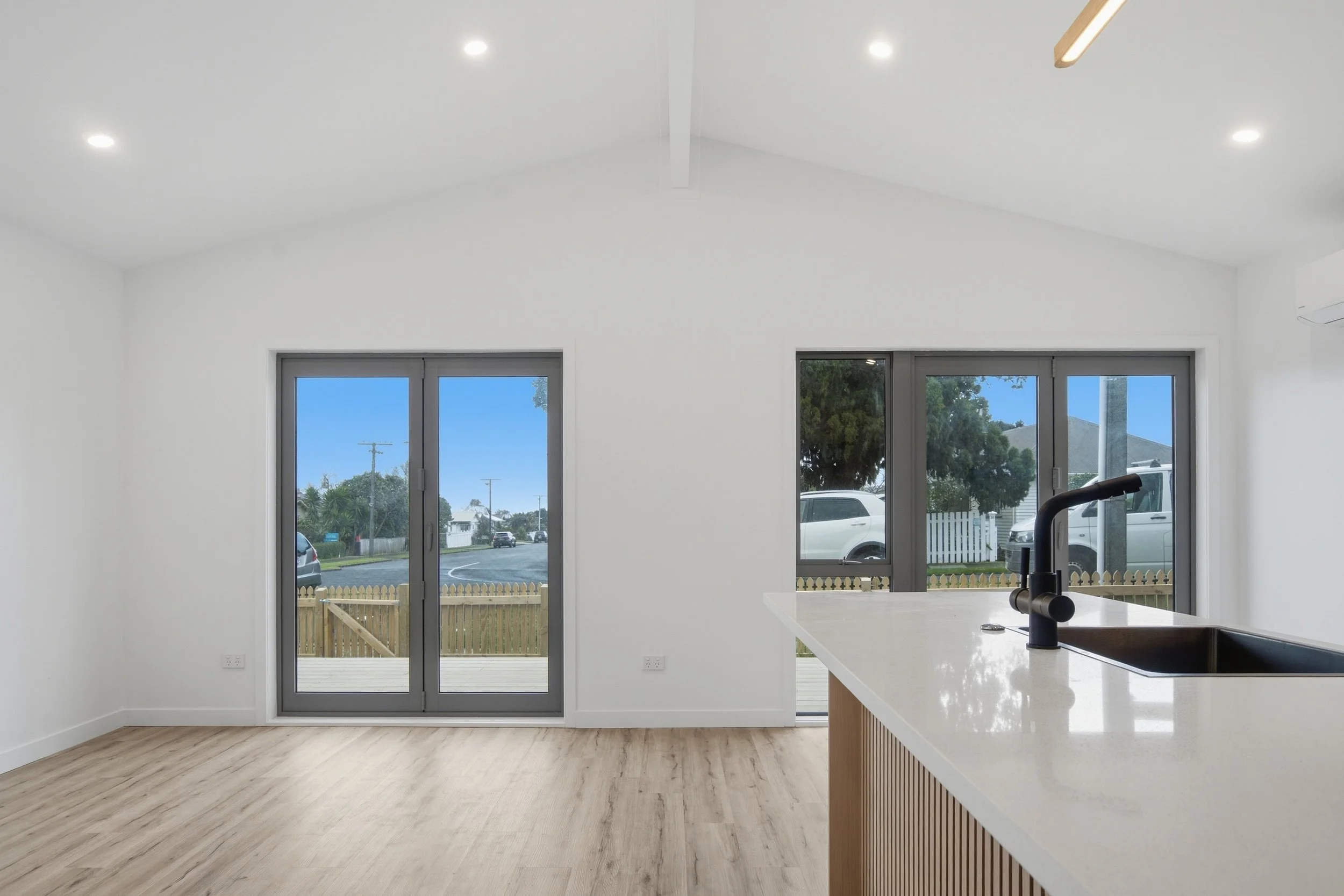Interior view of a modern, minimalist kitchen with white walls, a white countertop, a black faucet, and large windows showing a suburban neighborhood outside.