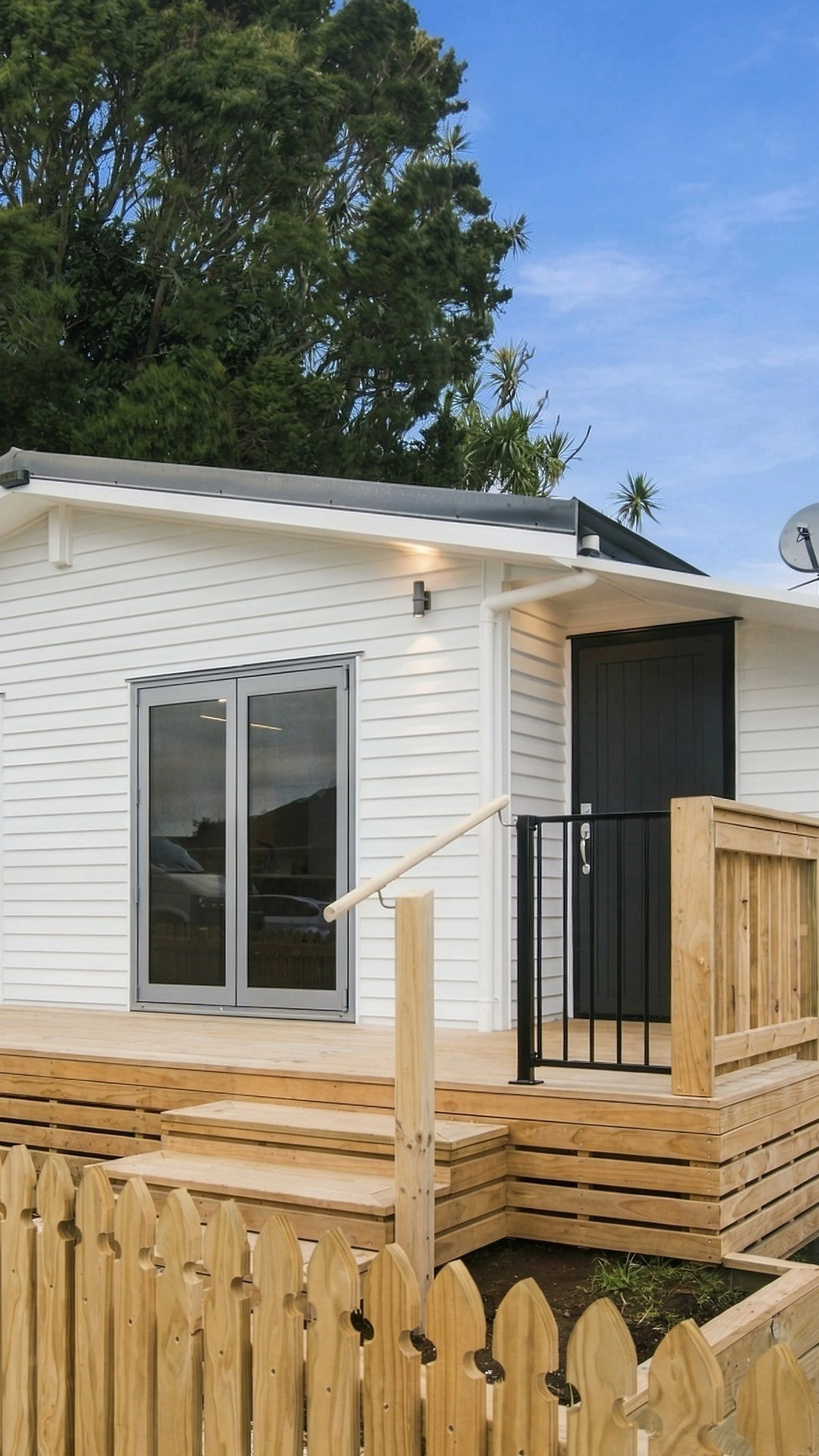 A white house with black doors and a front porch, surrounded by a wooden fence. There are trees in the background and a blue sky overhead.