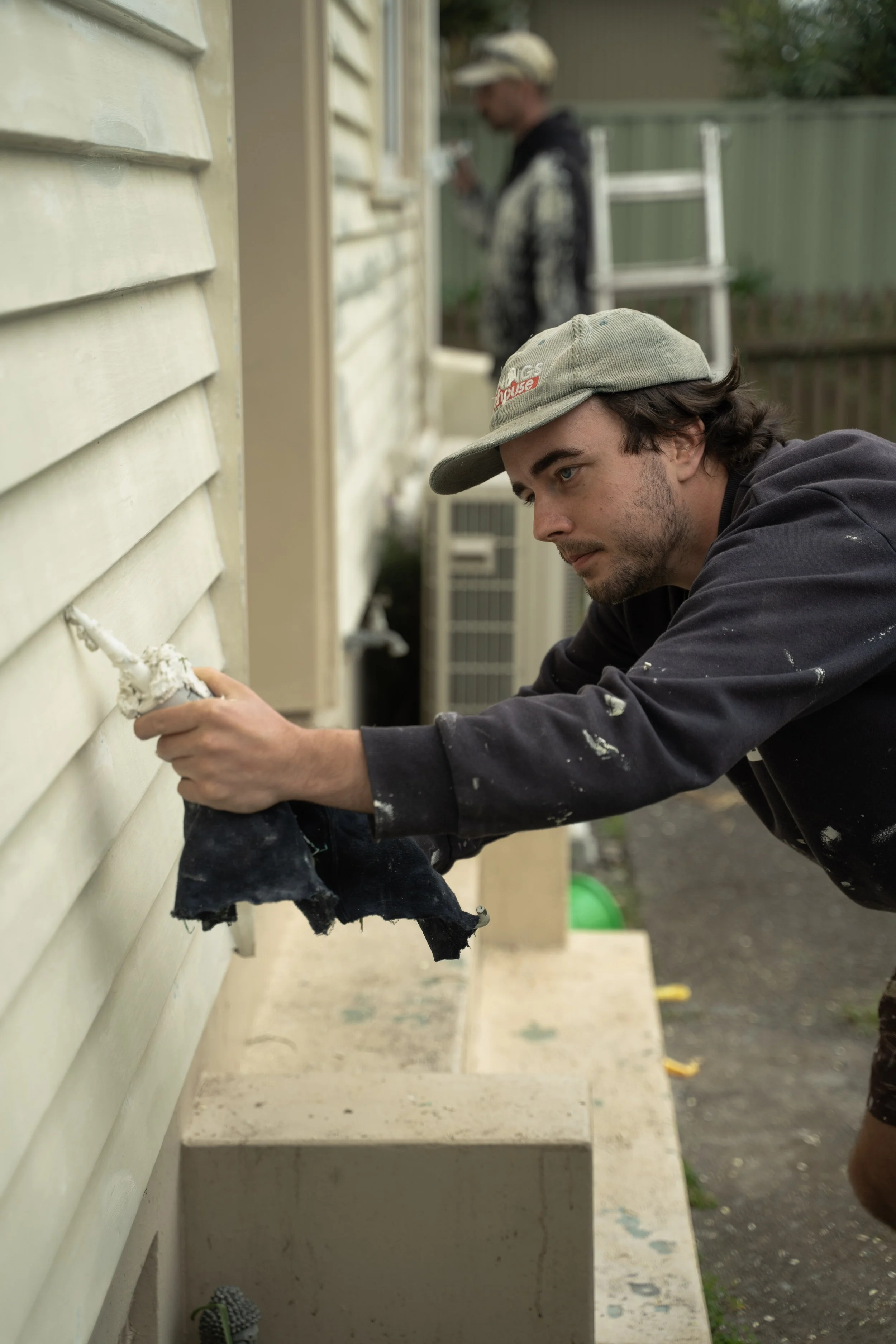 A man in a gray hoodie and cap is kneeling outside, applying white paint to the exterior siding of a house using a caulking gun. In the background, another man is working on a ladder.