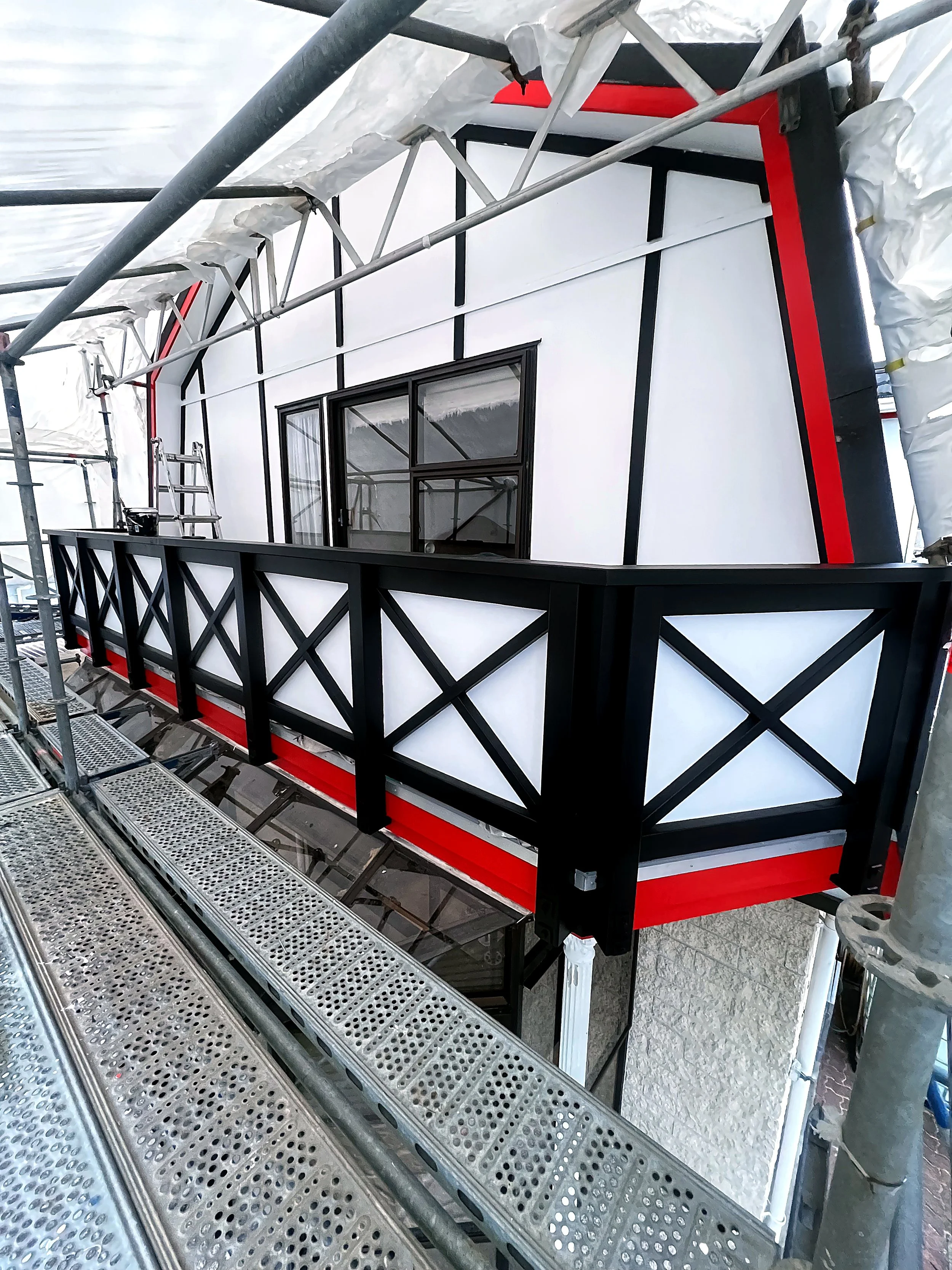 Close-up of a building under construction, showing scaffolding, a balcony with a black railing, and a window with black frames. The building has white walls with black and red trim, and construction materials and support structures are visible around the scene.