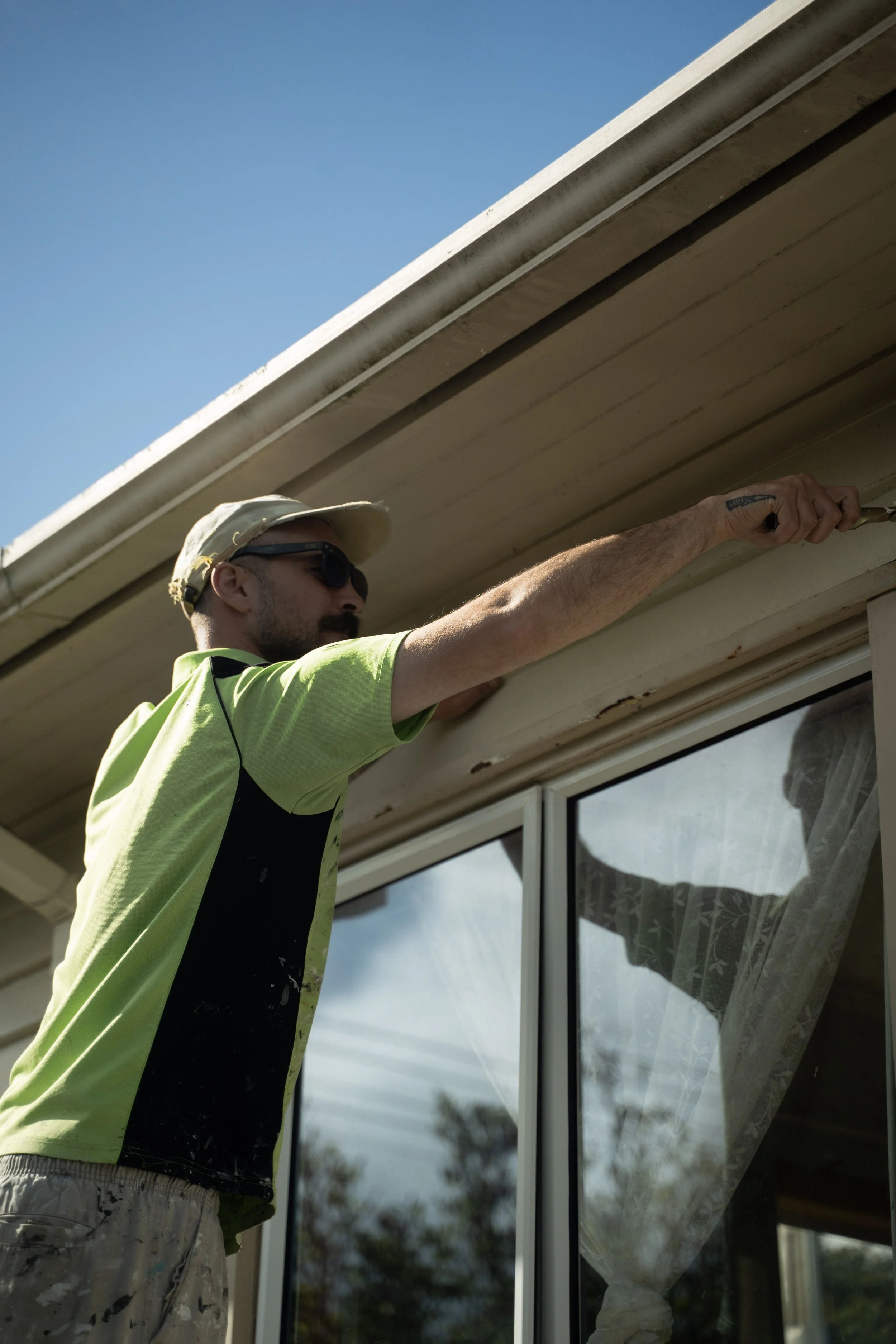 A man wearing sunglasses, a beige cap, a bright yellow-green high-visibility shirt, and paint-splattered pants is working on the exterior of a house. He is using a tool to either install or repair something under the eaves of the roof. The photo is taken outdoors, with trees and a blue sky in the background.