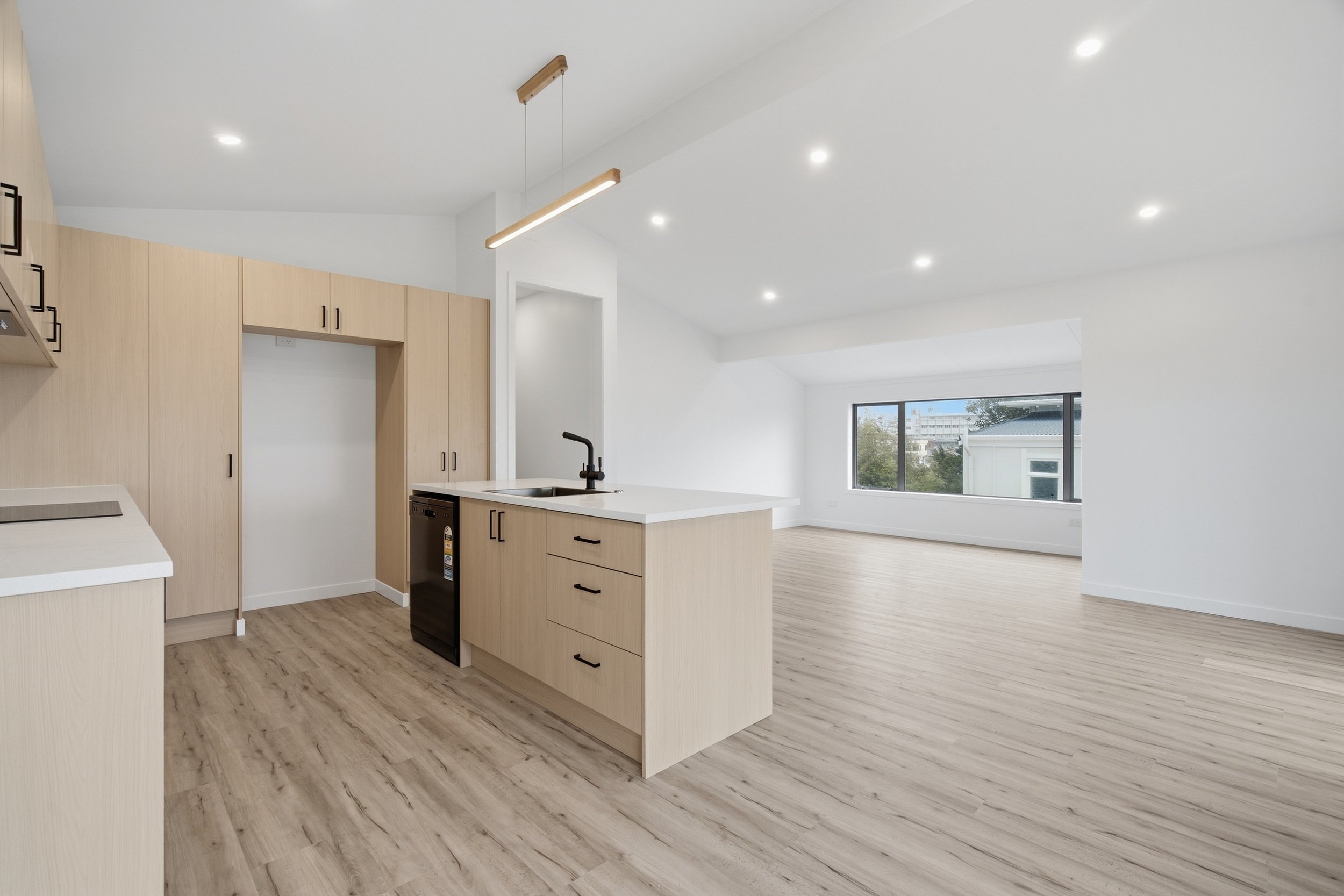 A modern kitchen with light wood cabinets, a kitchen island with a sink and black faucet, pendant lighting, and wood flooring, opening to a bright living space with a large window.