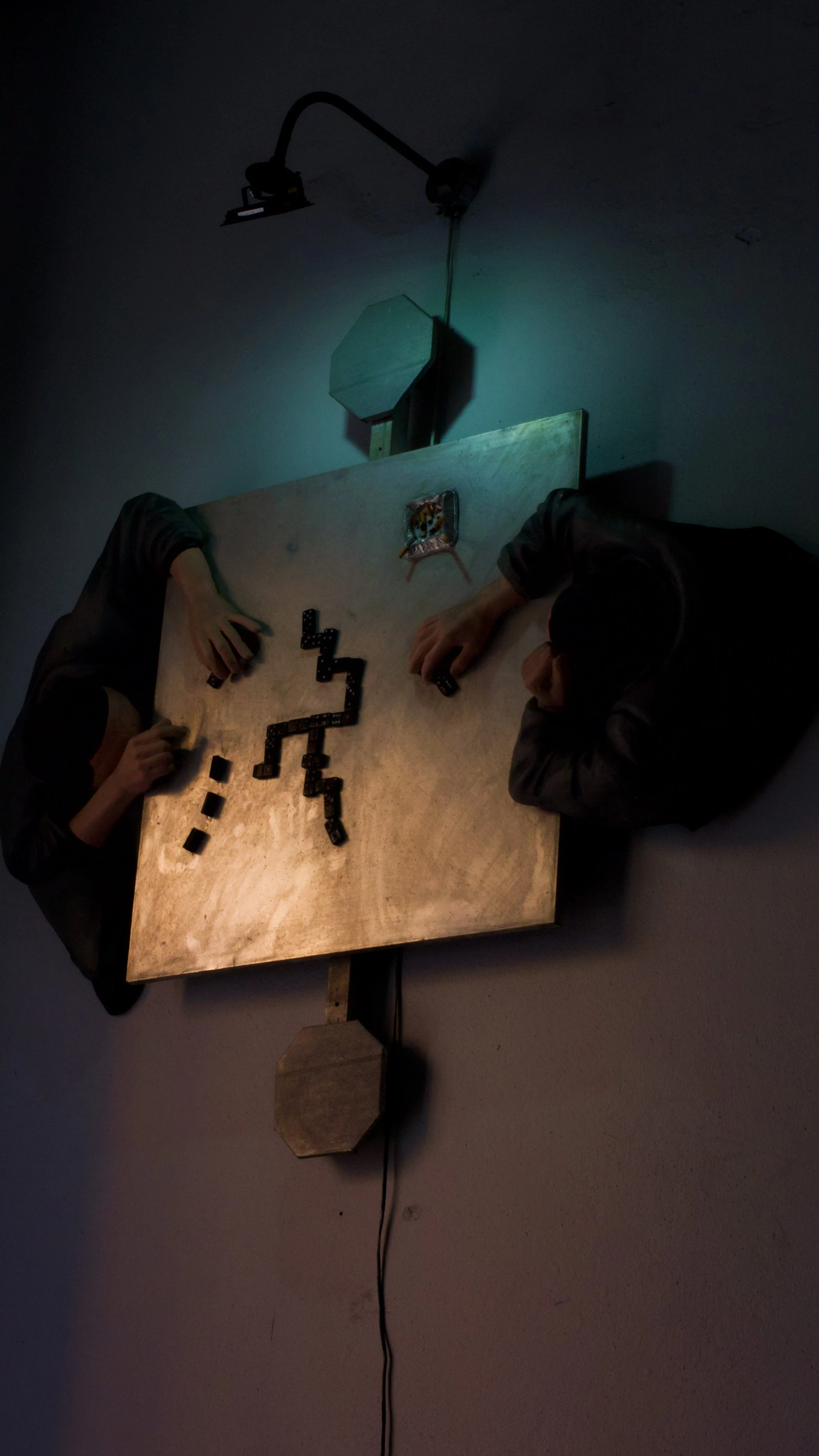 Two people are playing dominoes at a wooden table in a dimly lit room, viewed from above.