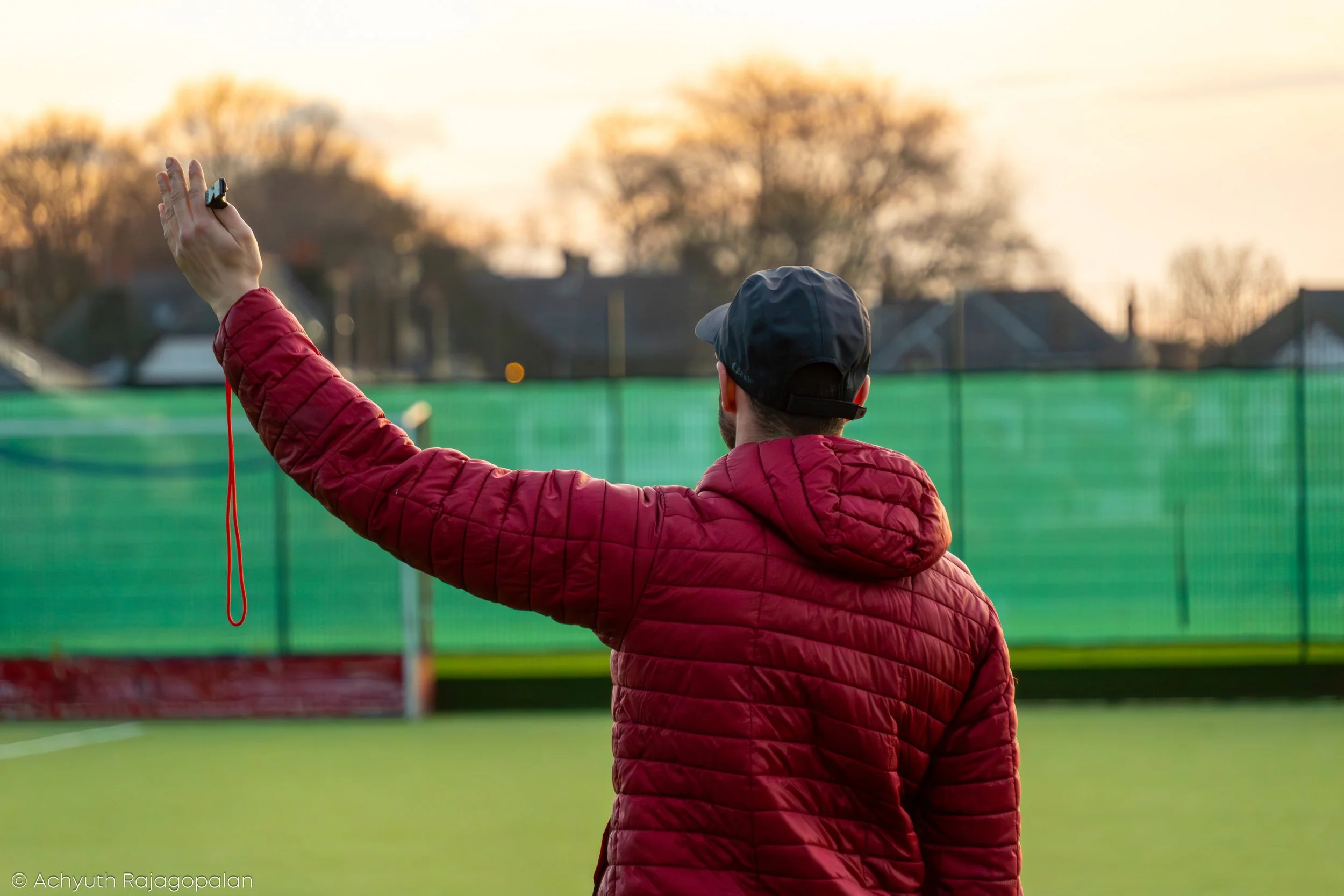 A person wearing a black cap and red quilted jacket is taking a selfie or photo with their phone outdoors during sunset, with trees and houses in the background.