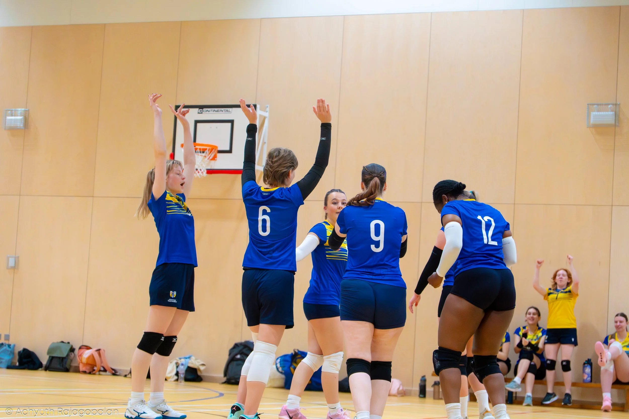 A group of female volleyball players in blue uniforms celebrate on an indoor court, some with their hands raised, with a basketball hoop and wooden wall in the background.