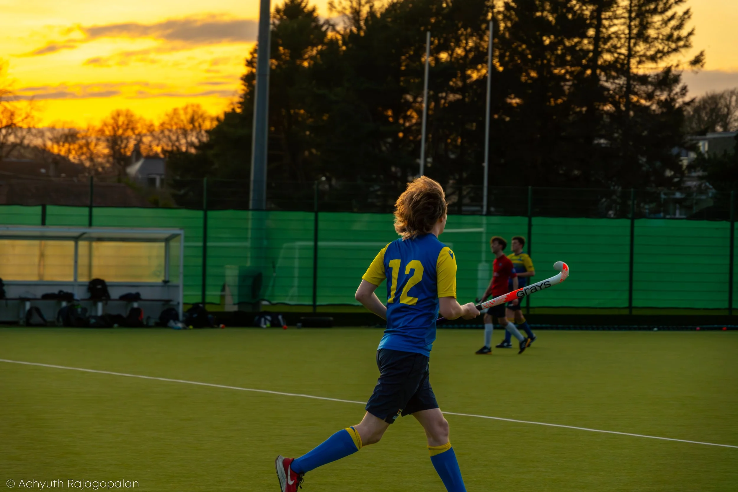 A young person with curly hair, wearing a blue and yellow sports jersey numbered 12, running on a field during sunset with a hockey stick in hand and other players in the background.