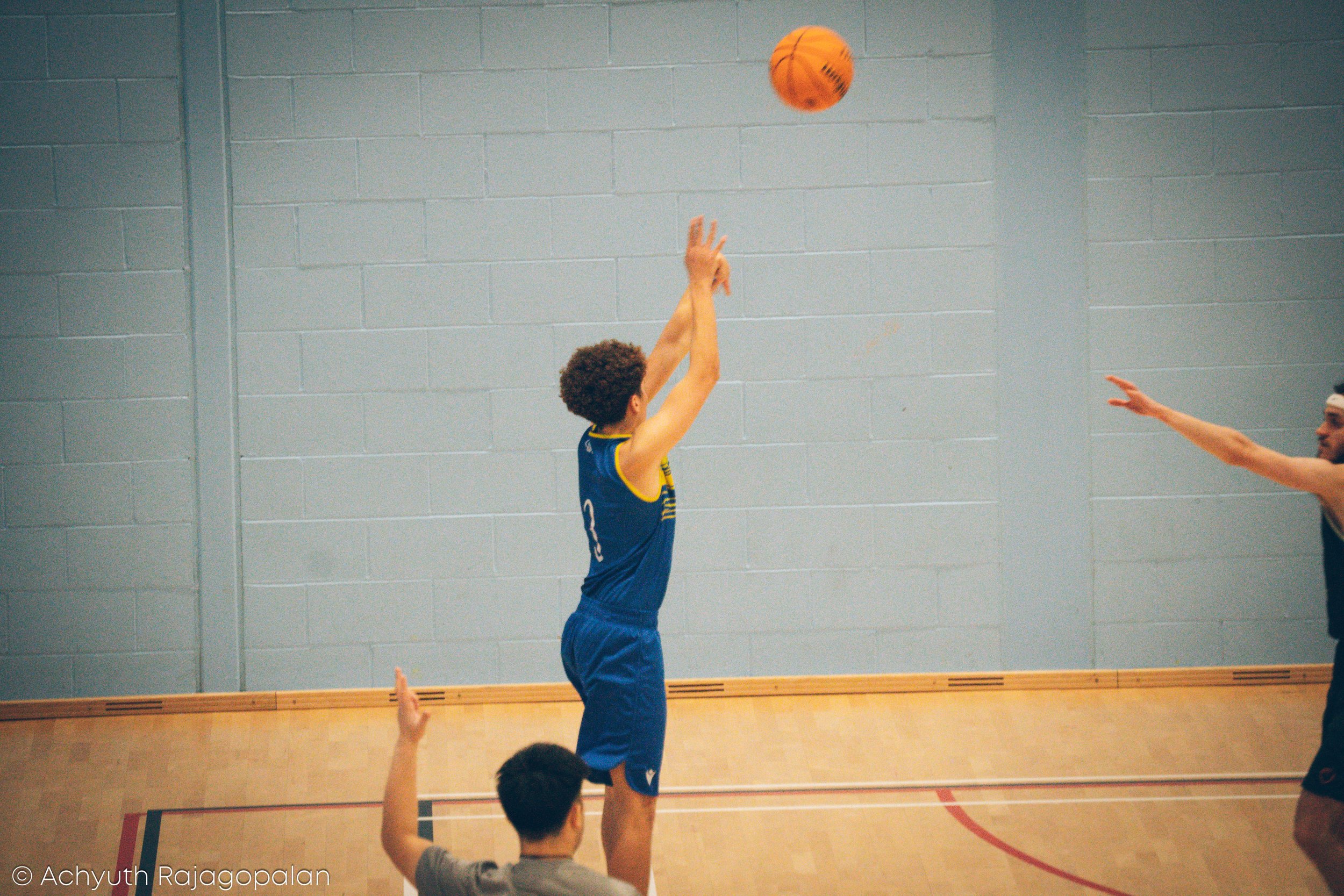 A person in a blue sports jersey is playing basketball, jumping to shoot or pass the ball while another person extends an arm in defense or block. The setting appears to be an indoor gymnasium with a gray brick wall and wooden floor.