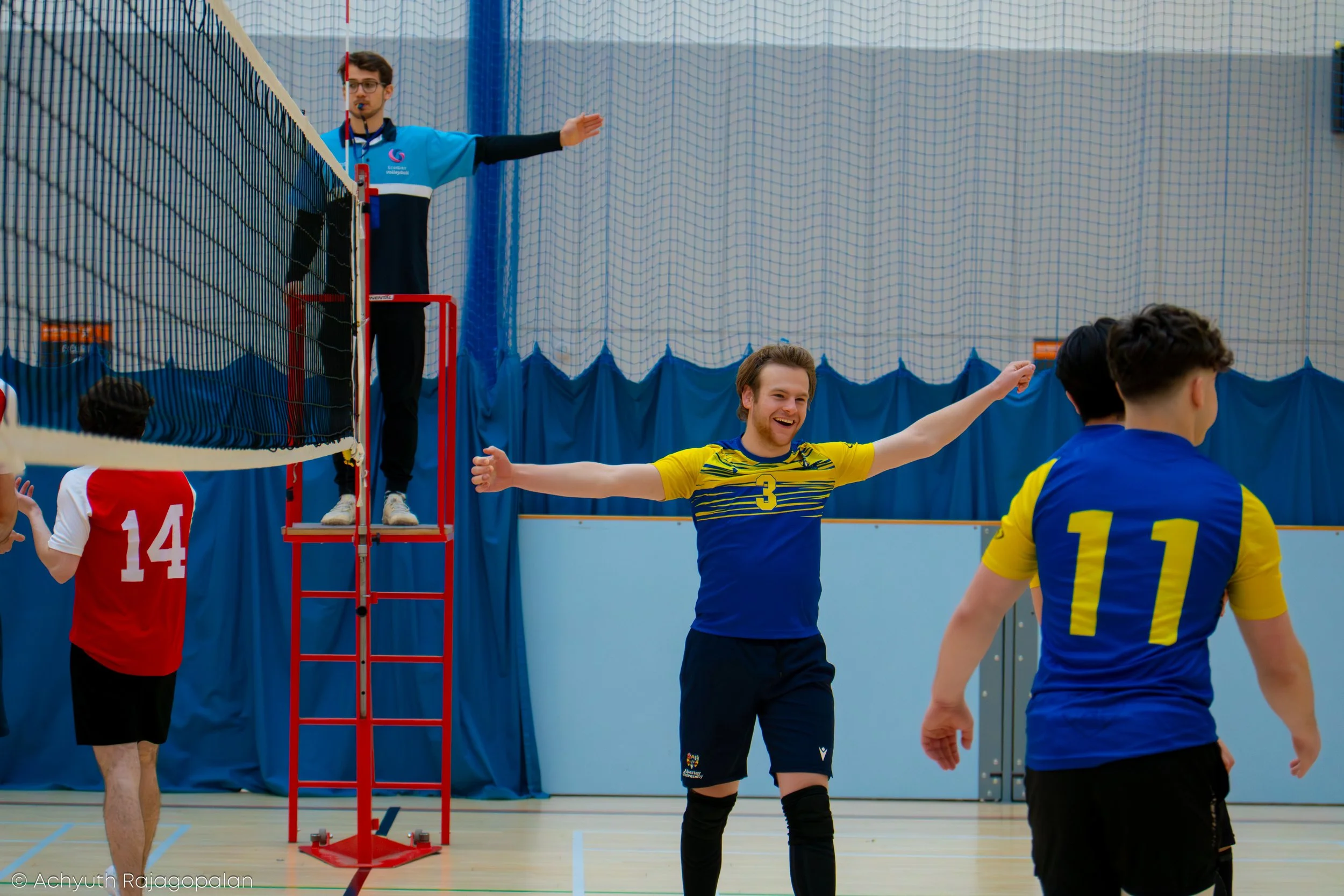 Indoor volleyball game with players celebrating a point, a referee on a stand, and a net with blue padding, blue curtains in the background.