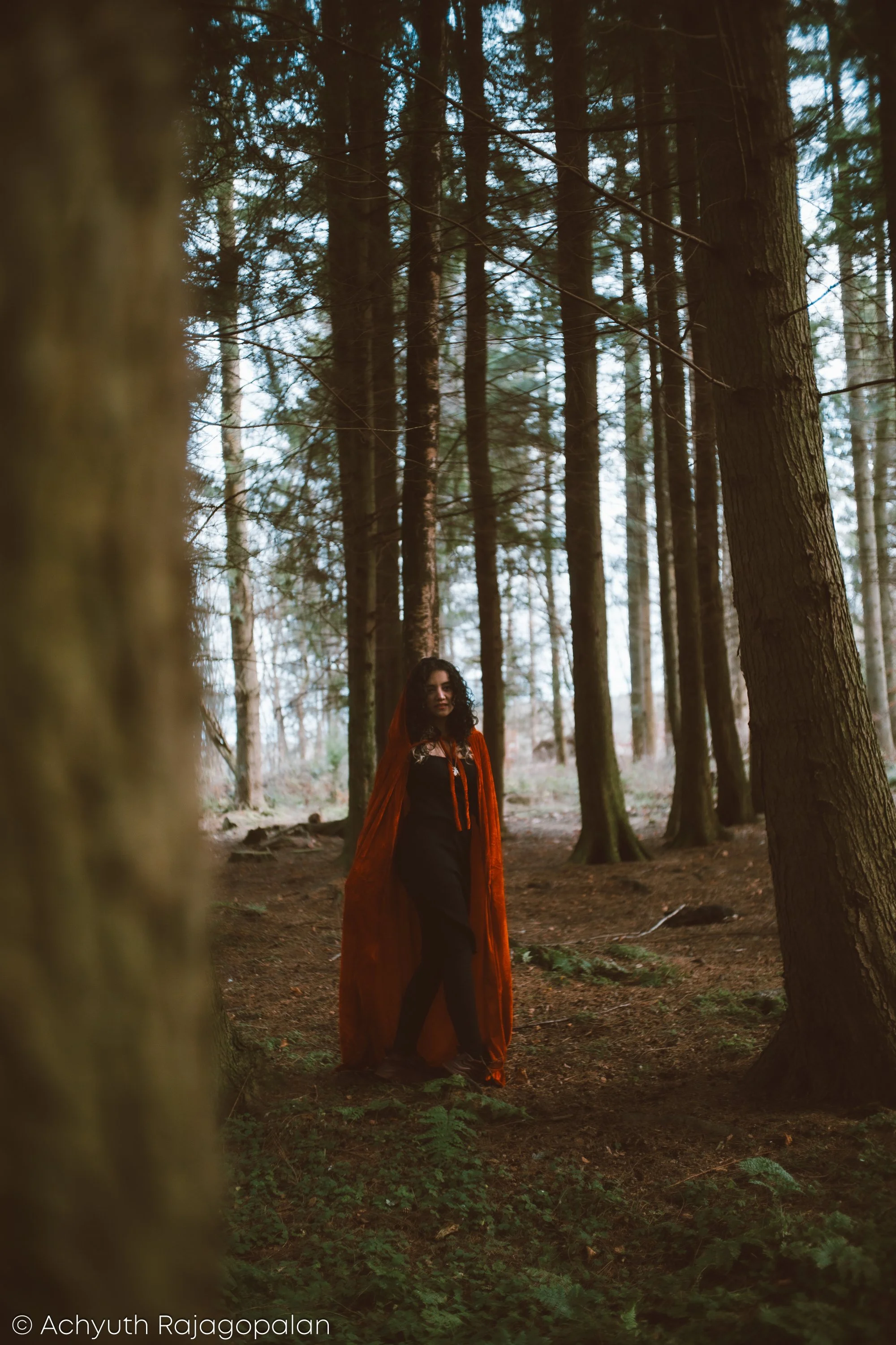 A woman with dark curly hair and light skin in a forest with tall trees, wearing a black dress and a flowing orange cape.