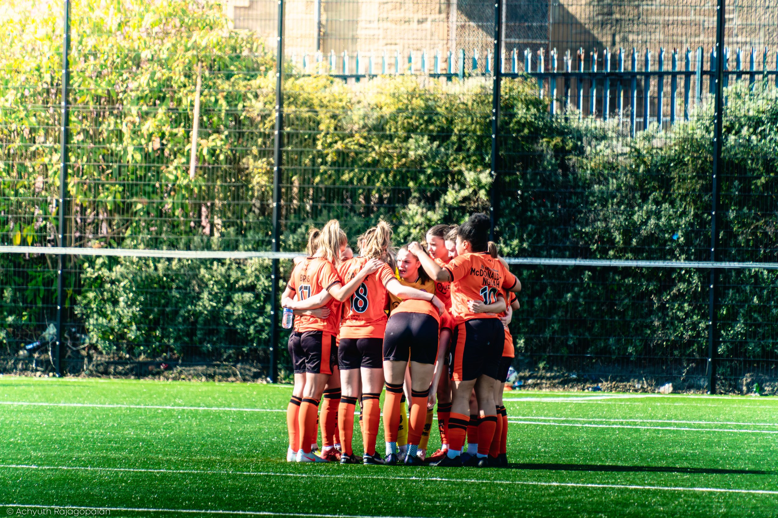 A group of female soccer players wearing orange uniforms huddle together on a soccer field.