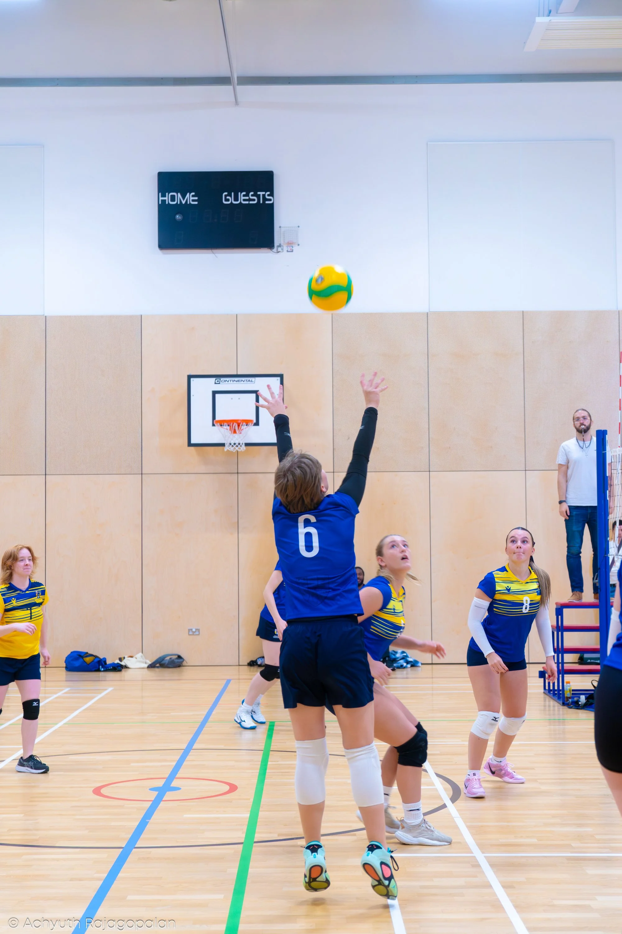 A volleyball game in progress inside a gymnasium. A player in a blue jersey with the number 6 on the back is jumping to hit the ball, while other players observe and prepare. The gym has a wooden floor, a basketball hoop, and a scoreboard on the wall