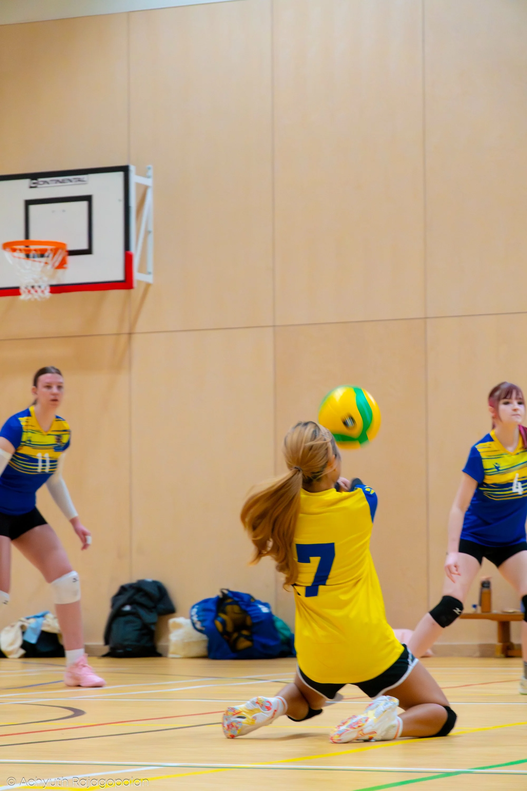 Girls playing volleyball in an indoor gym, one girl in yellow jersey number 7 is on the floor in a diving position, others are standing and watching.