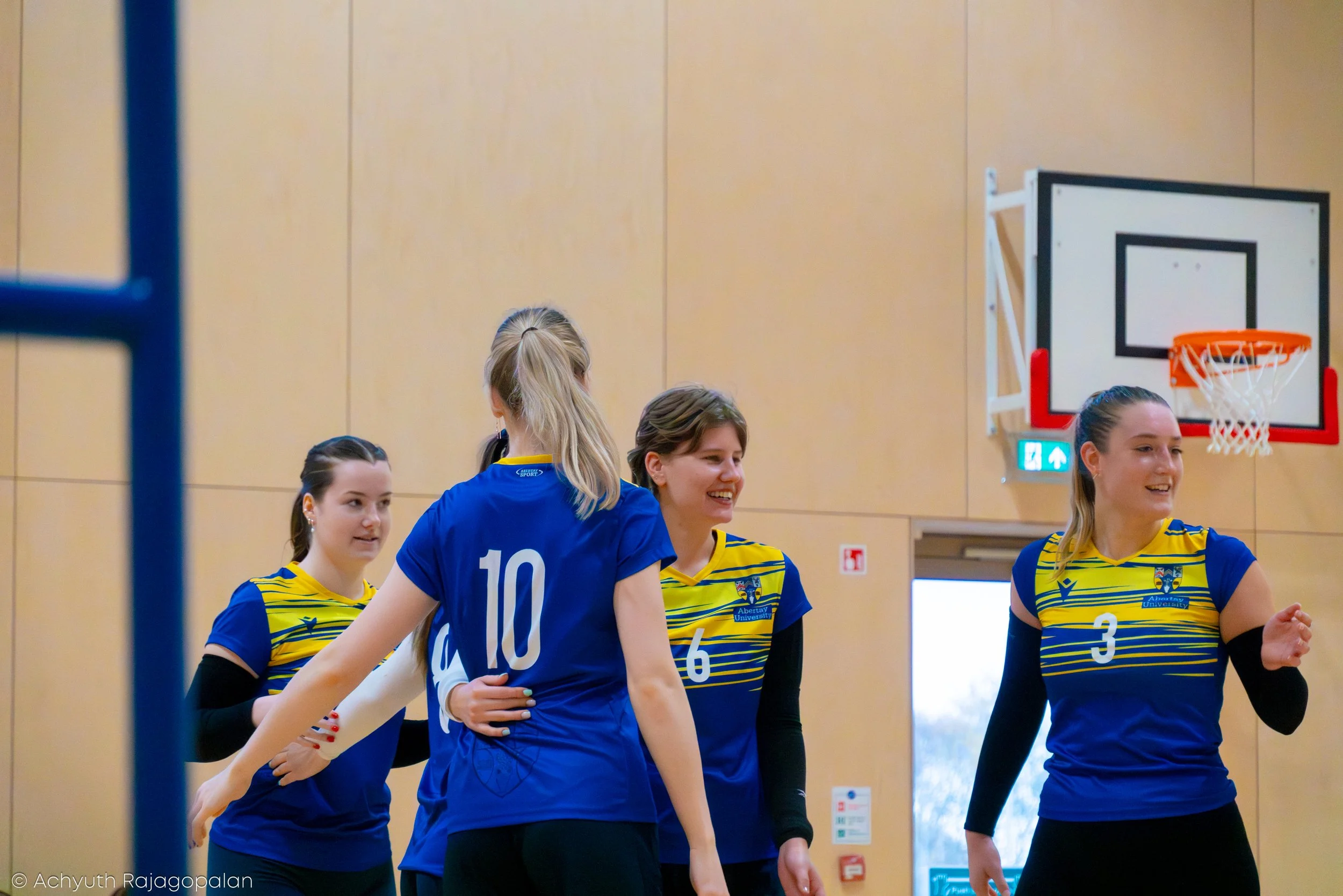 A group of female volleyball players on an indoor court, wearing yellow and blue uniforms, standing together and smiling. One player is wearing a dark blue jersey with the number 10 on the back.