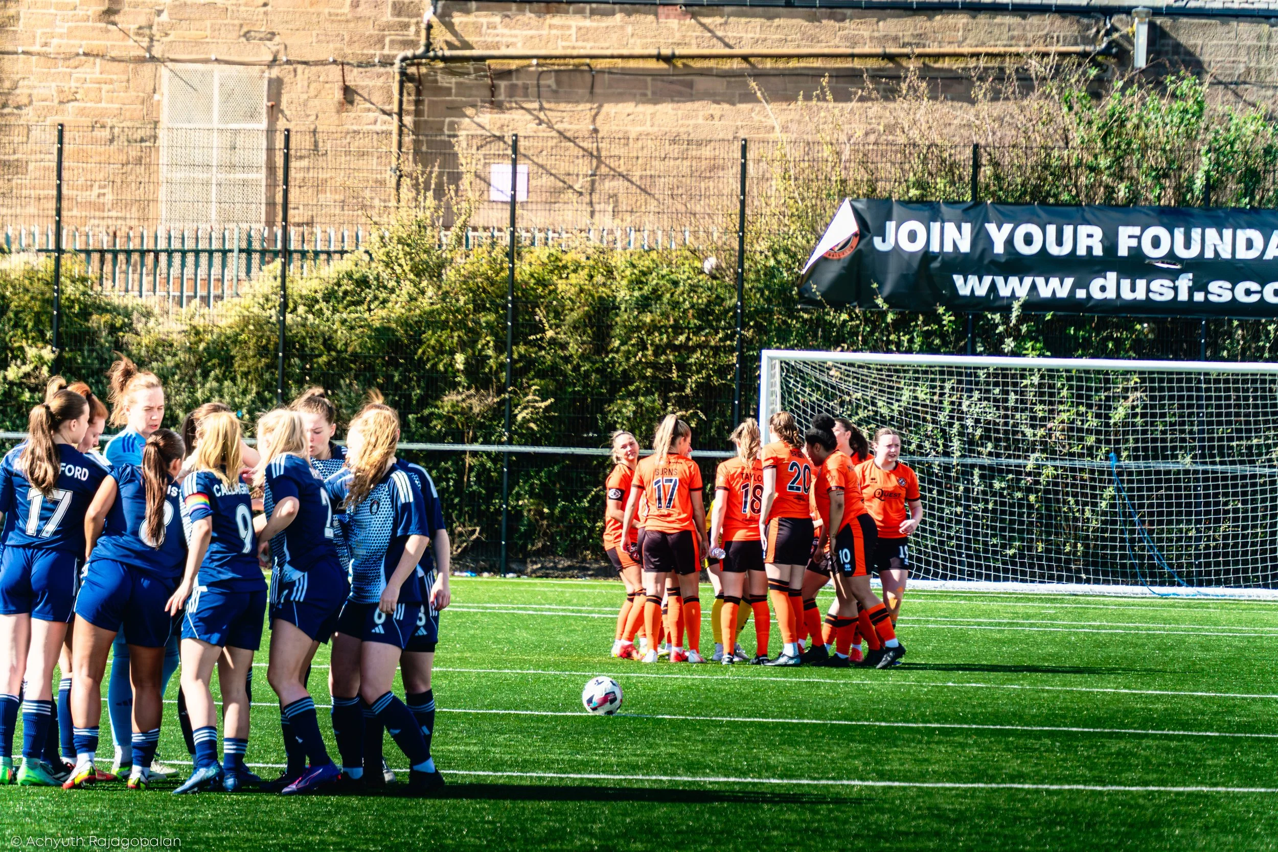 Young women soccer players in blue and orange jerseys standing on a soccer field, facing each other, with a soccer ball on the ground and a goalpost in the background.