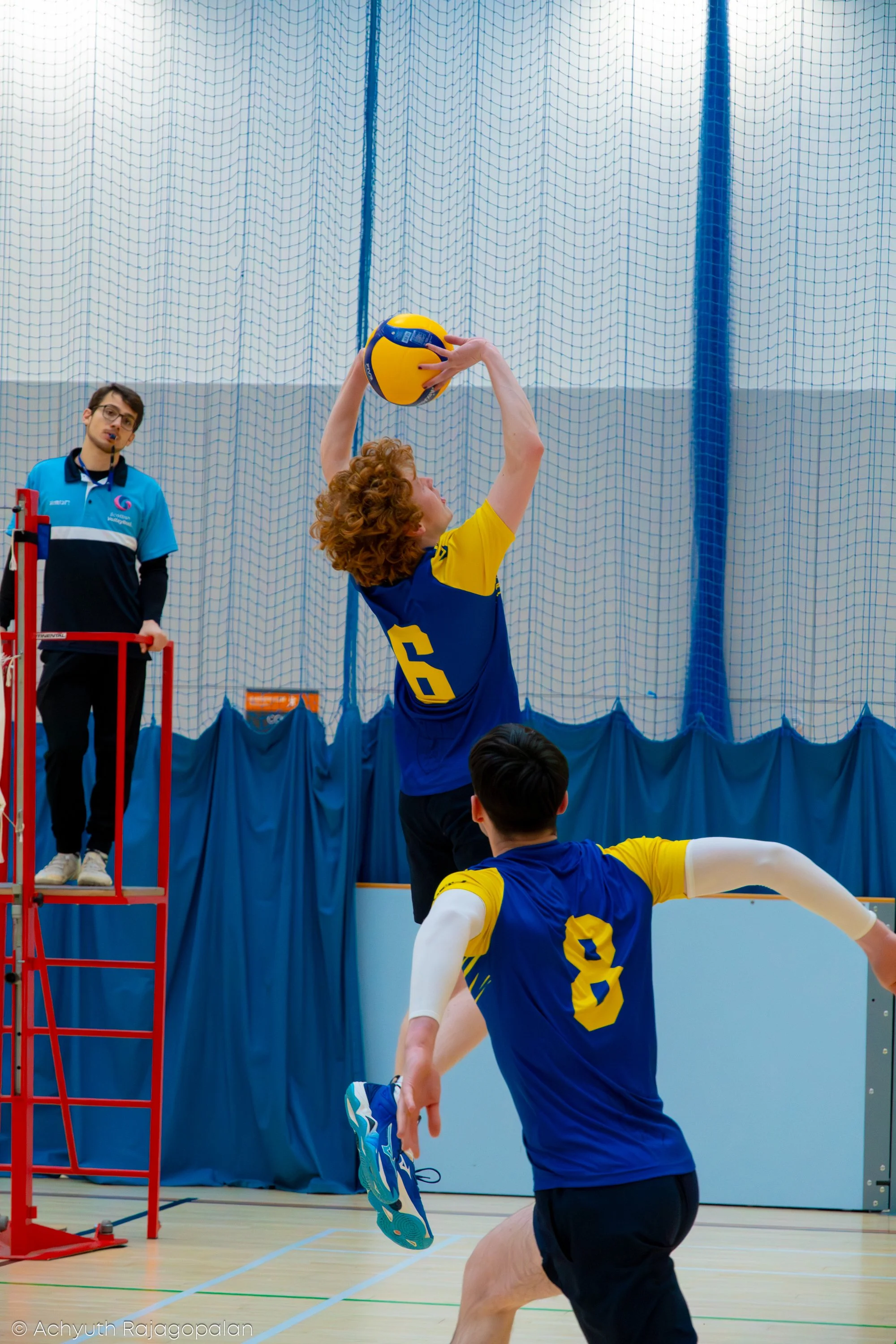 A volleyball player with curly red hair dressed in a blue and yellow uniform is jumping to hit a volleyball in an indoor volleyball court. Another player, wearing a similar uniform, is preparing to catch or block, while a referee or coach stands near