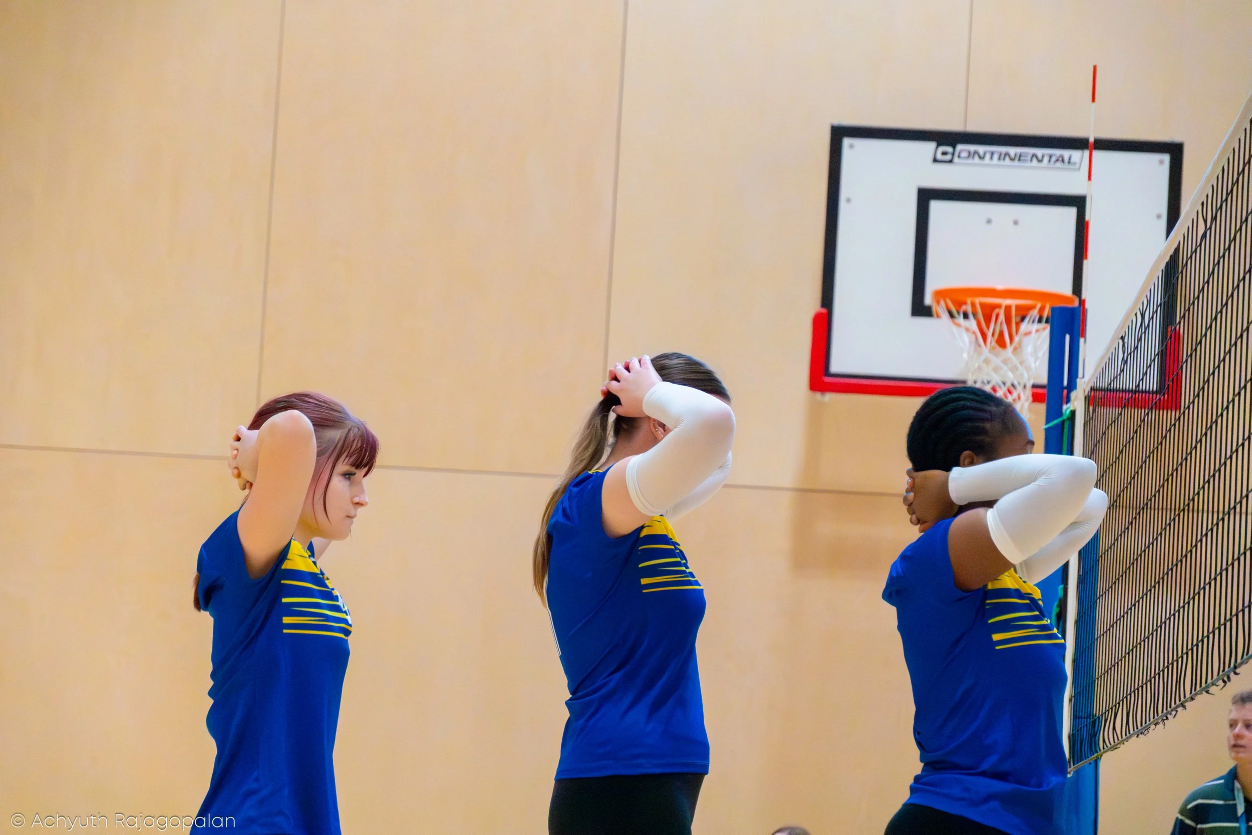 Group of young female volleyball players standing with hands on their heads during a game in an indoor gym, with a basketball hoop visible on the wall.