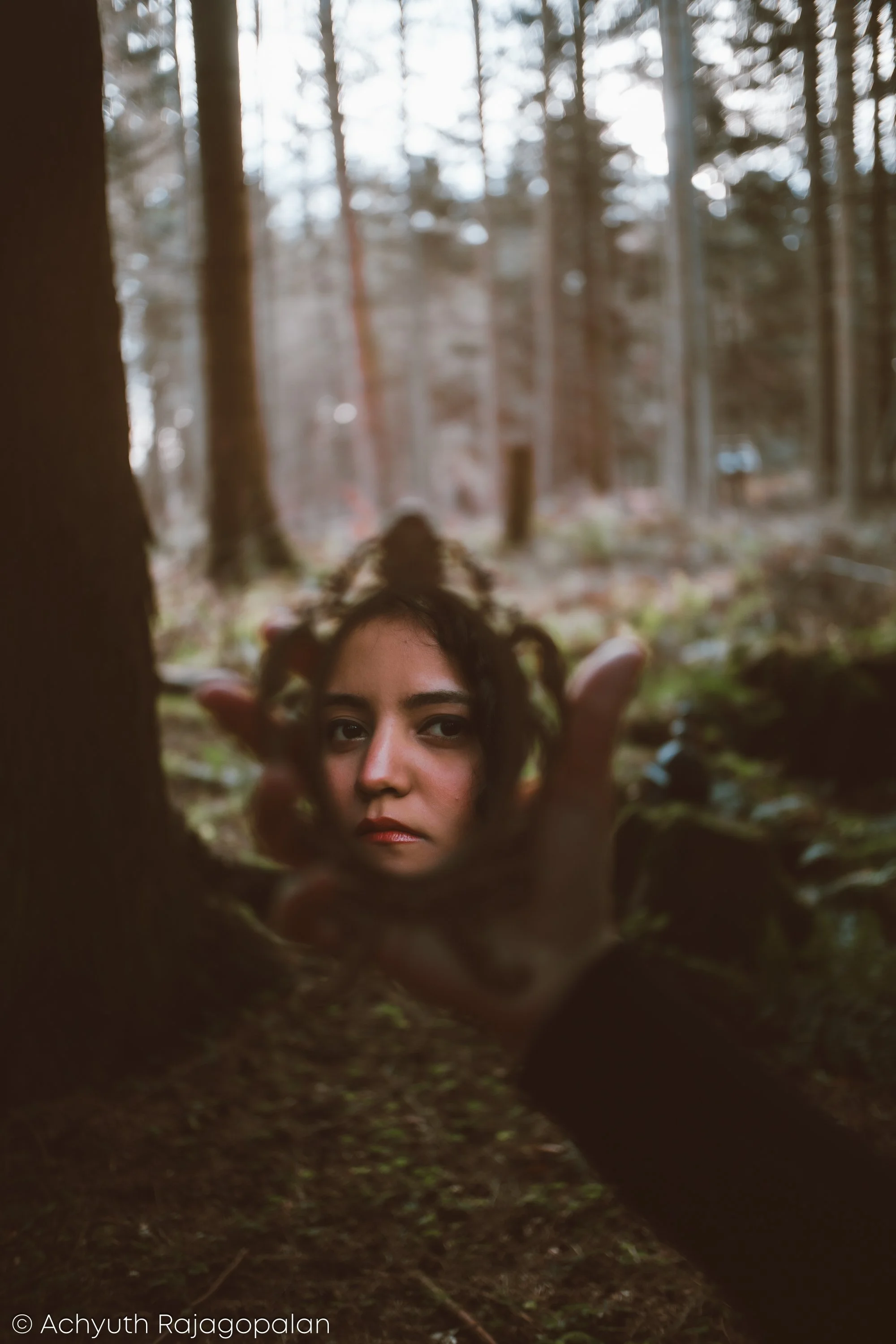 A woman with dark curly hair peeking through a small, circular opening in a tree trunk in a forest.